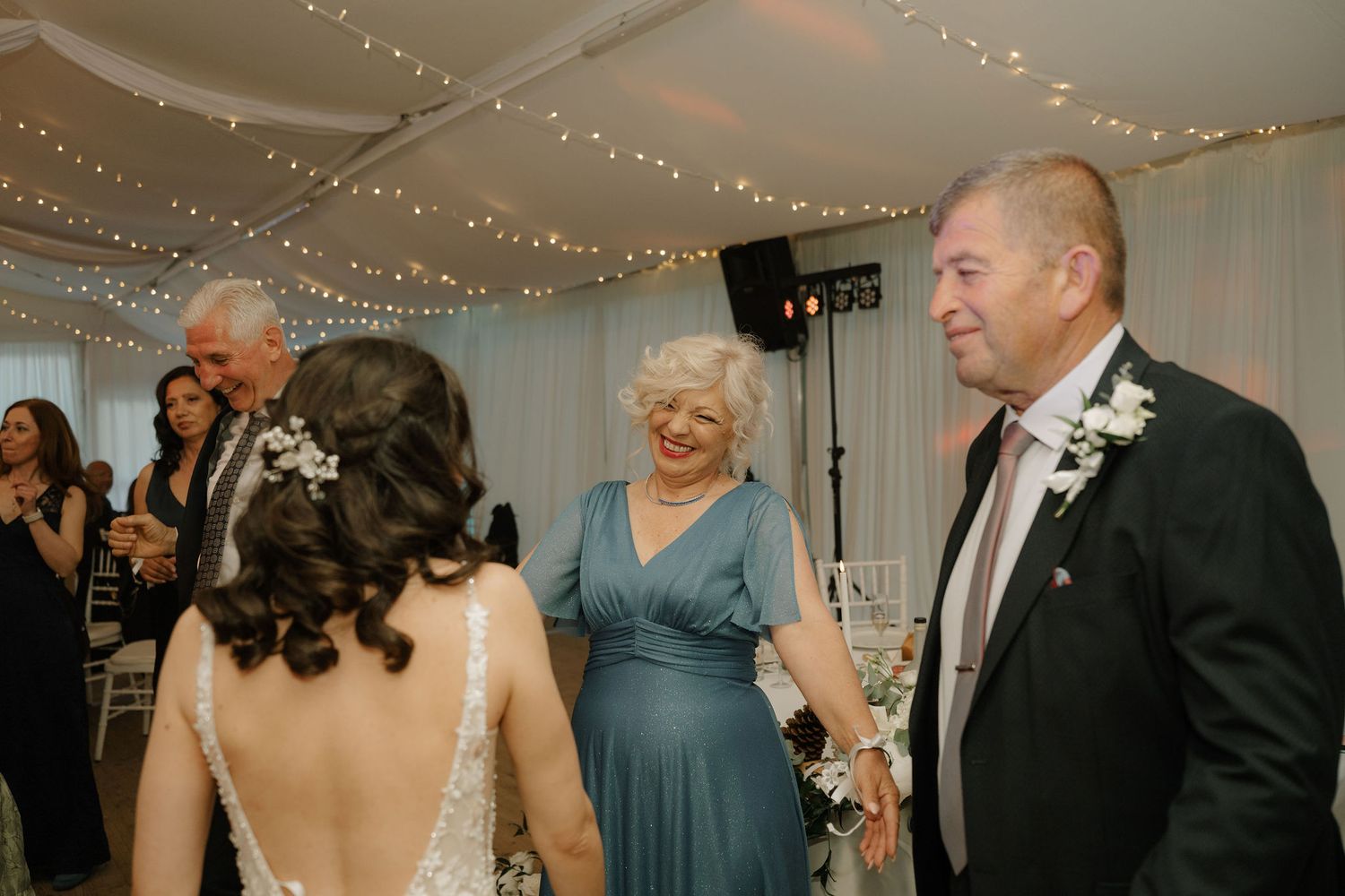Bride in lace gown dancing with smiling elderly couple at elegant wedding reception with string lights.