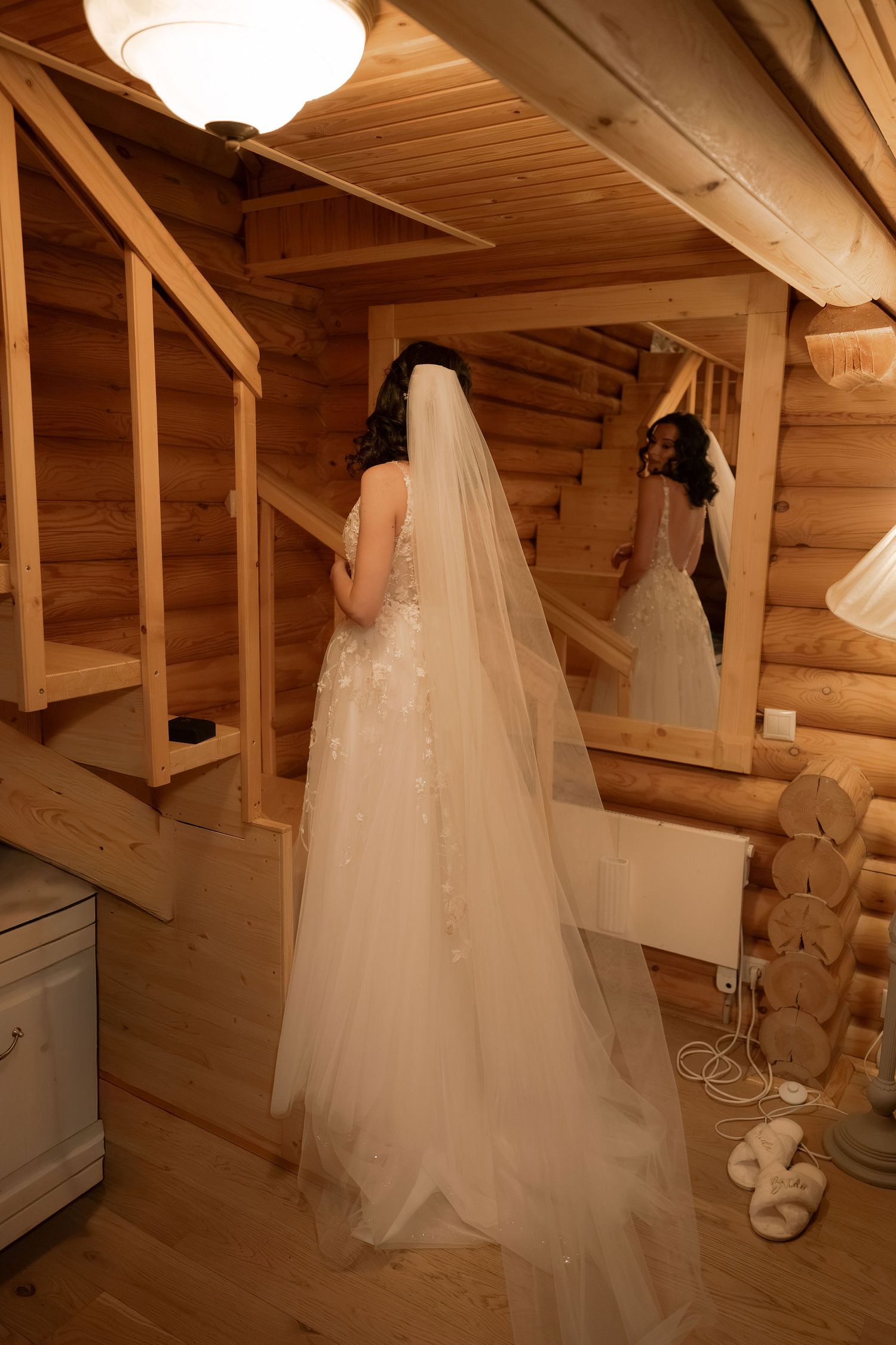 Bride in white wedding gown and veil stands before mirror in rustic log cabin interior.
