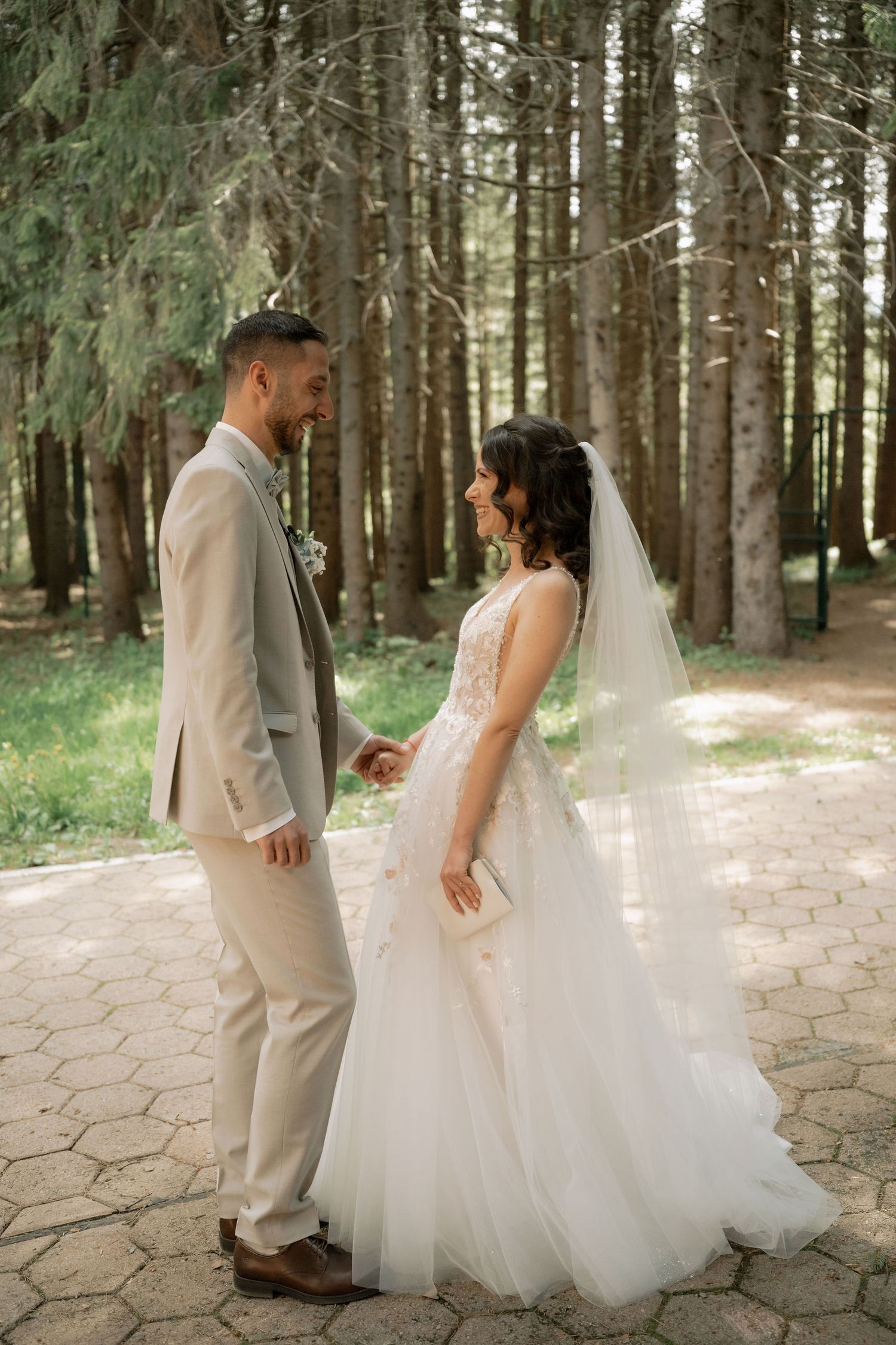A bride and groom hold hands and smile at each other in a forest setting, she in a white gown and he in a beige suit.