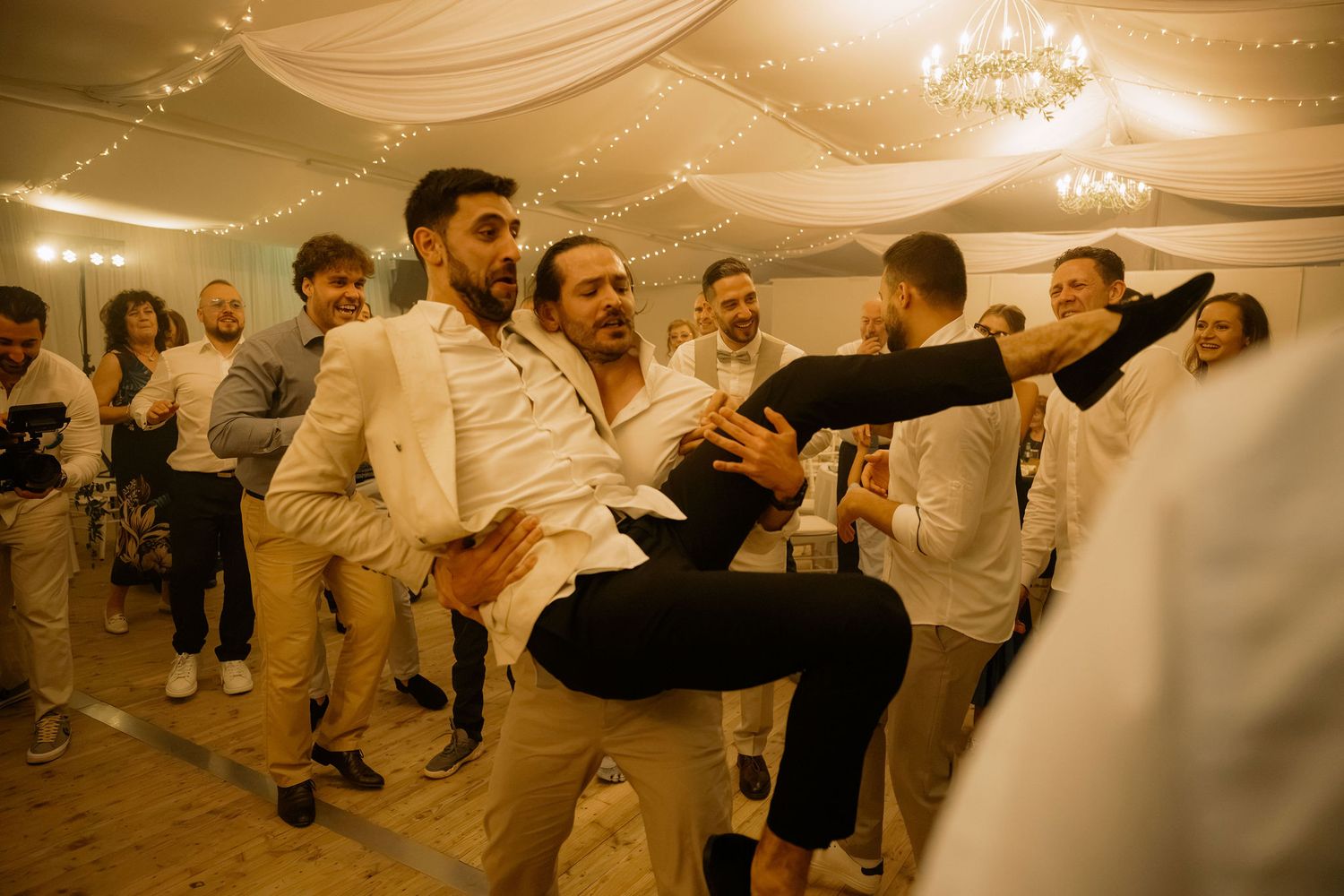 Guests dancing energetically at a wedding reception in a decorated tent venue with warm string lights.