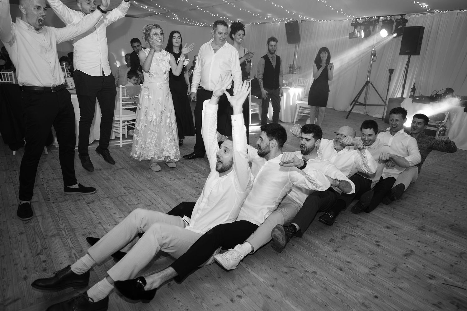 Black and white wedding reception photo of groosmen performing a train dance on the floor while guests cheer.