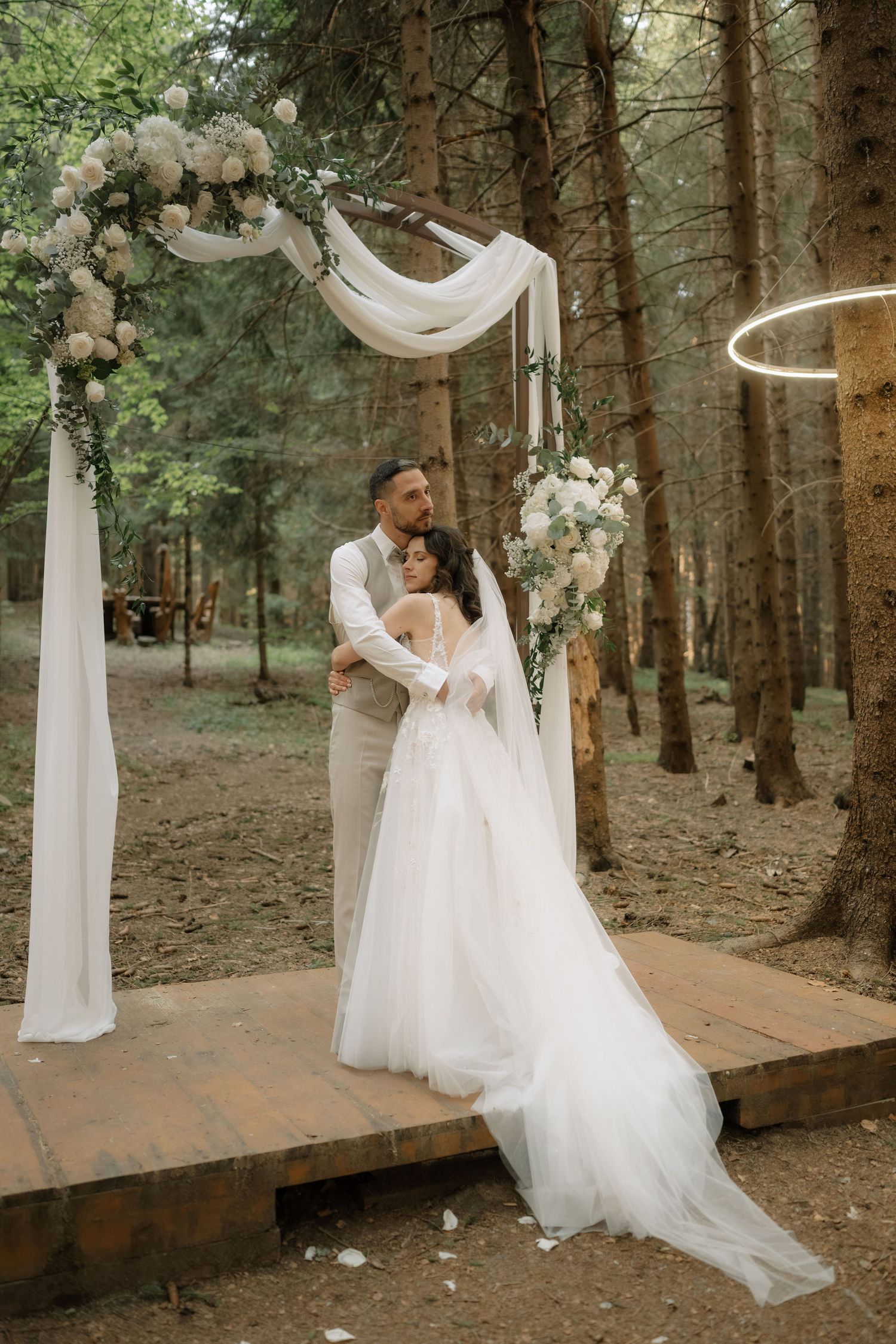 Bride and groom embrace under a floral arch with white draping in a forest wedding ceremony.