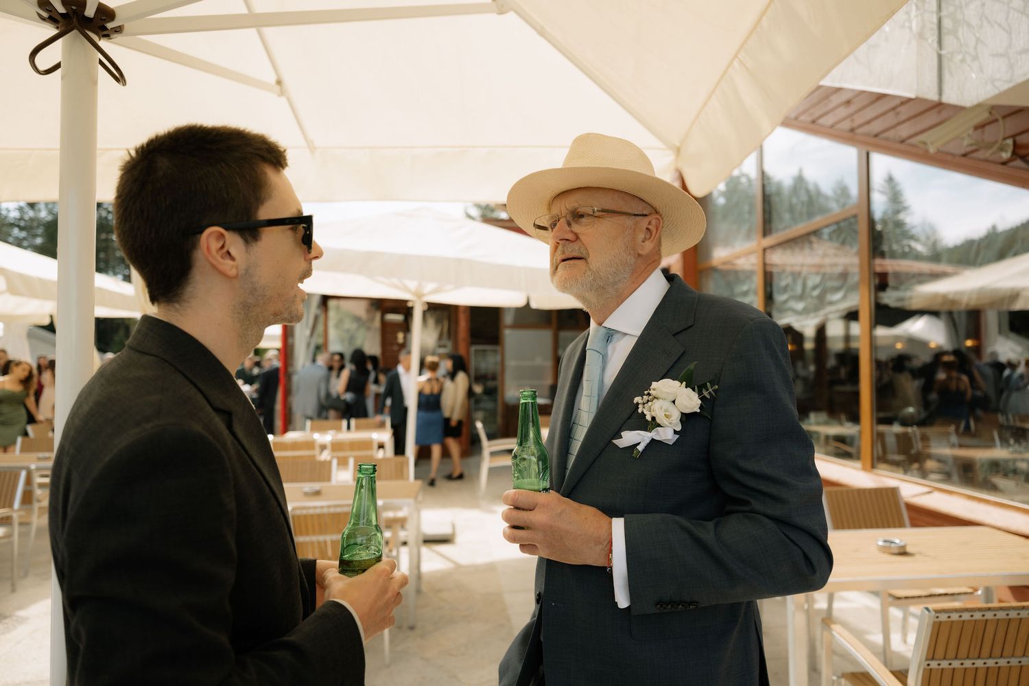 Two men in formal attire converse outdoors at a wedding reception, one wearing a cowboy hat and boutonniere.