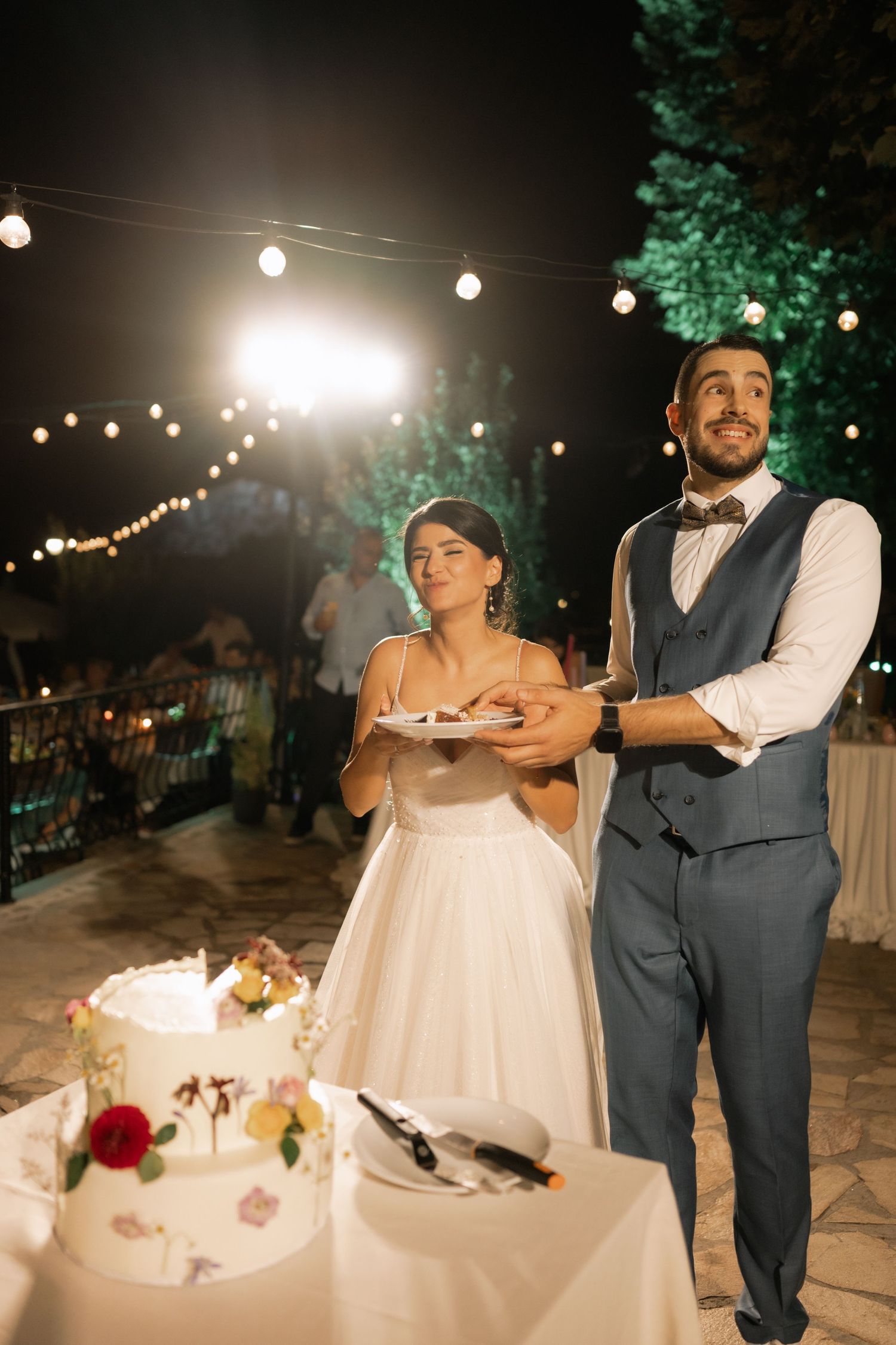 A couple in wedding attire shares a romantic moment under string lights during their outdoor evening reception celebration.
