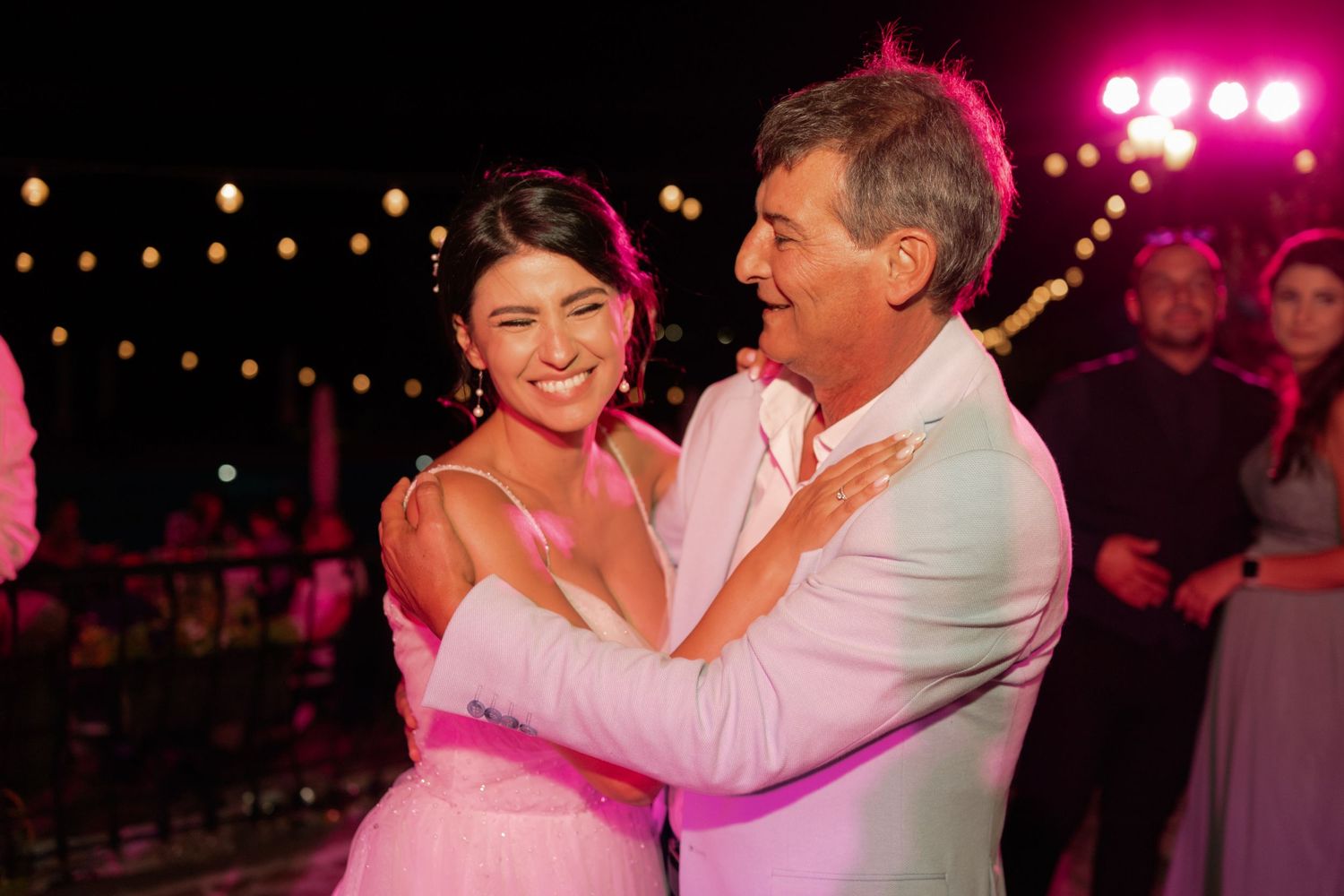 A couple dances together at an evening reception with pink lighting and string lights overhead.