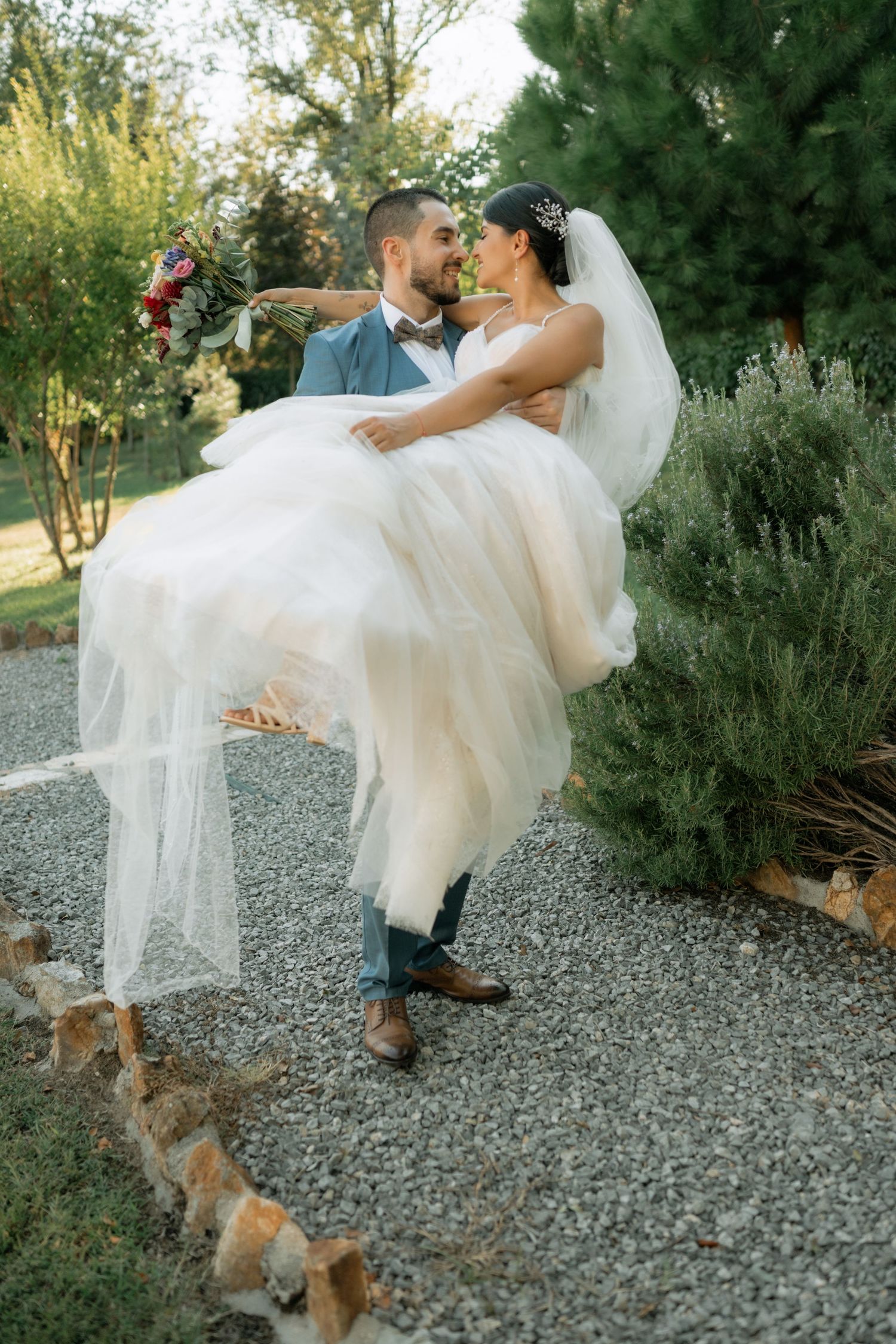 A couple in wedding attire shares a romantic moment on a gravel path while their dog watches nearby.