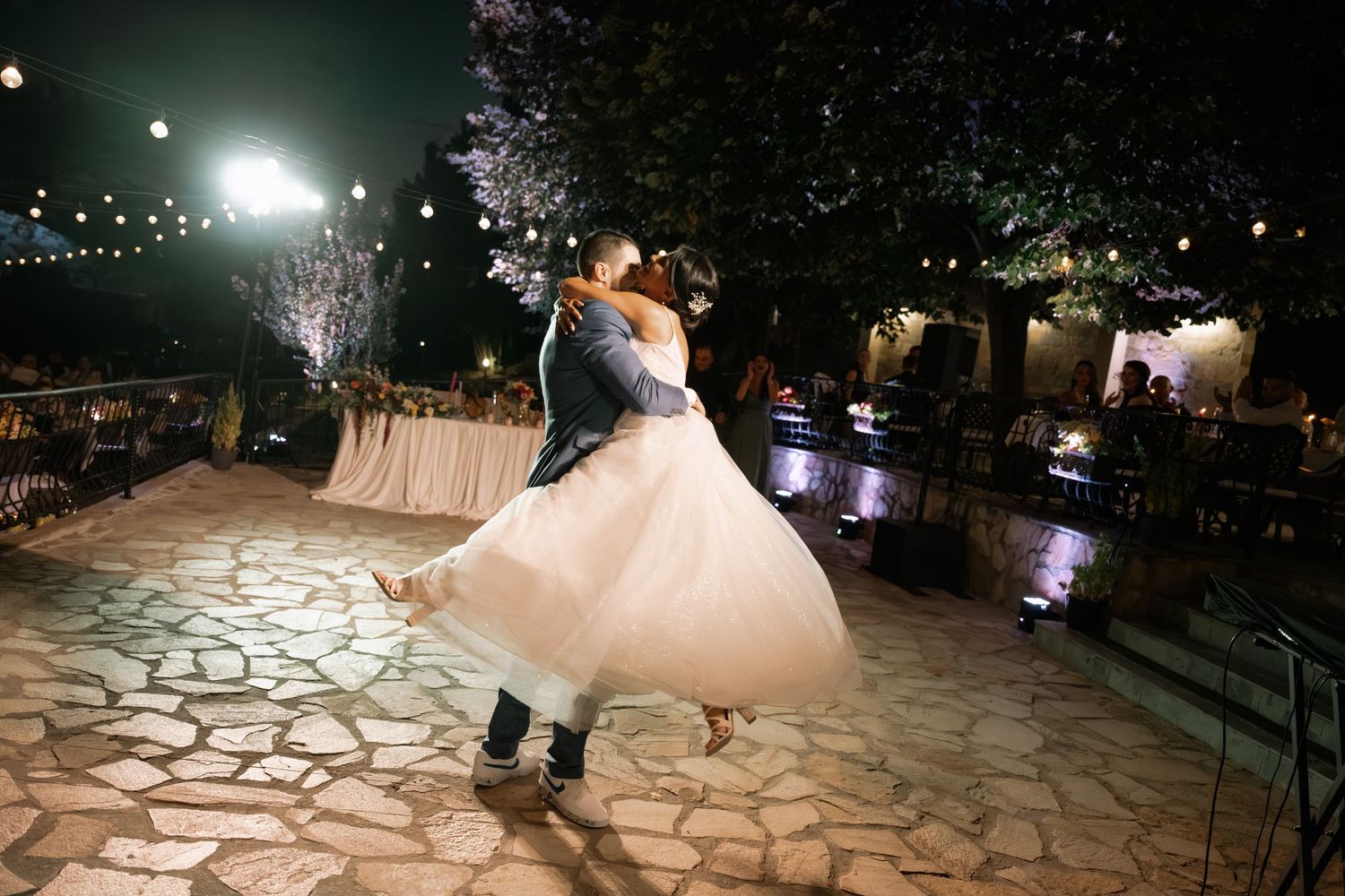 A bride in a white ballgown twirls on a stone patio at night with illuminated trees in the background.
