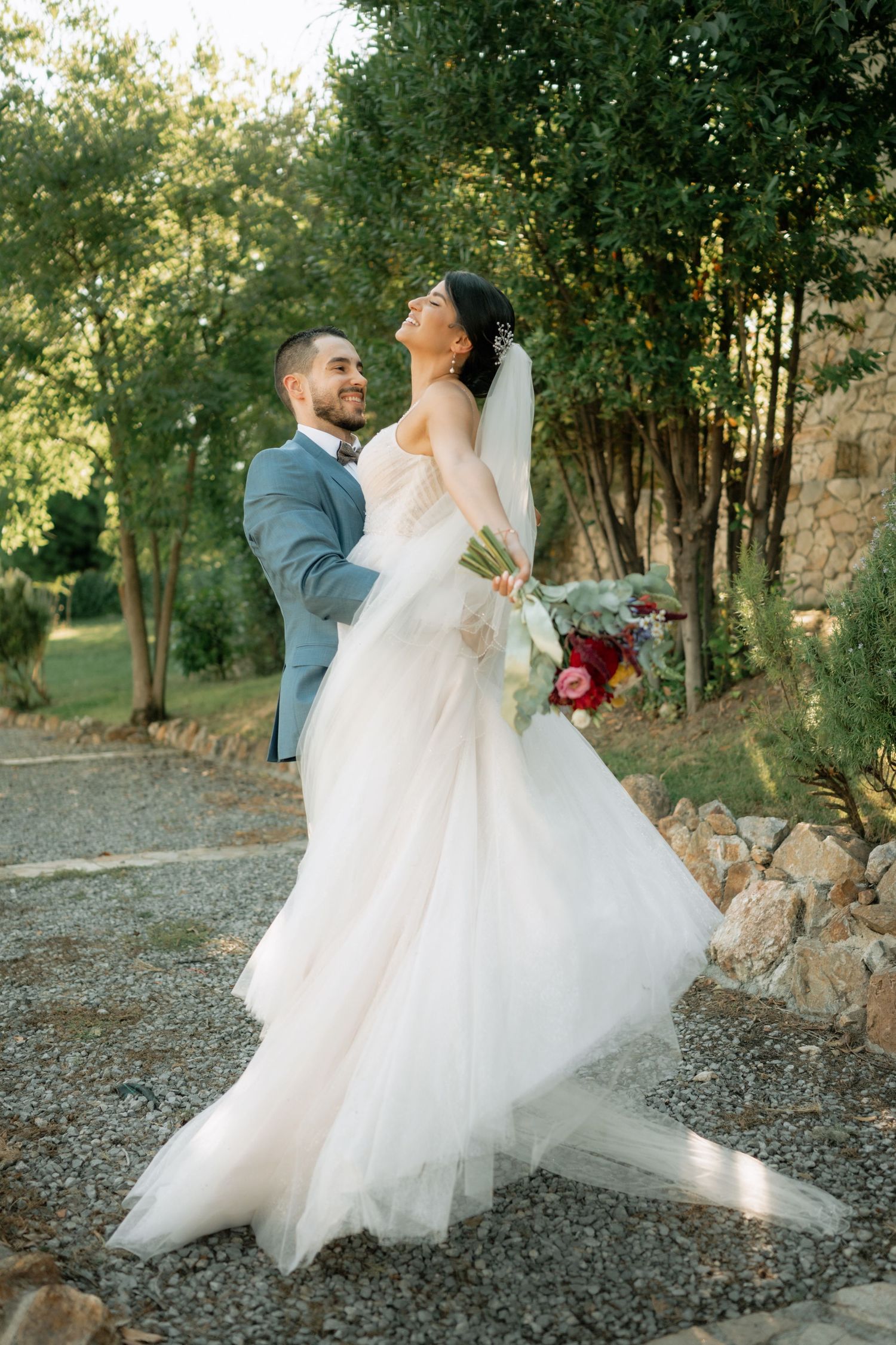 A joyful wedding couple shares a candid moment together on a gravel path surrounded by greenery at sunset.