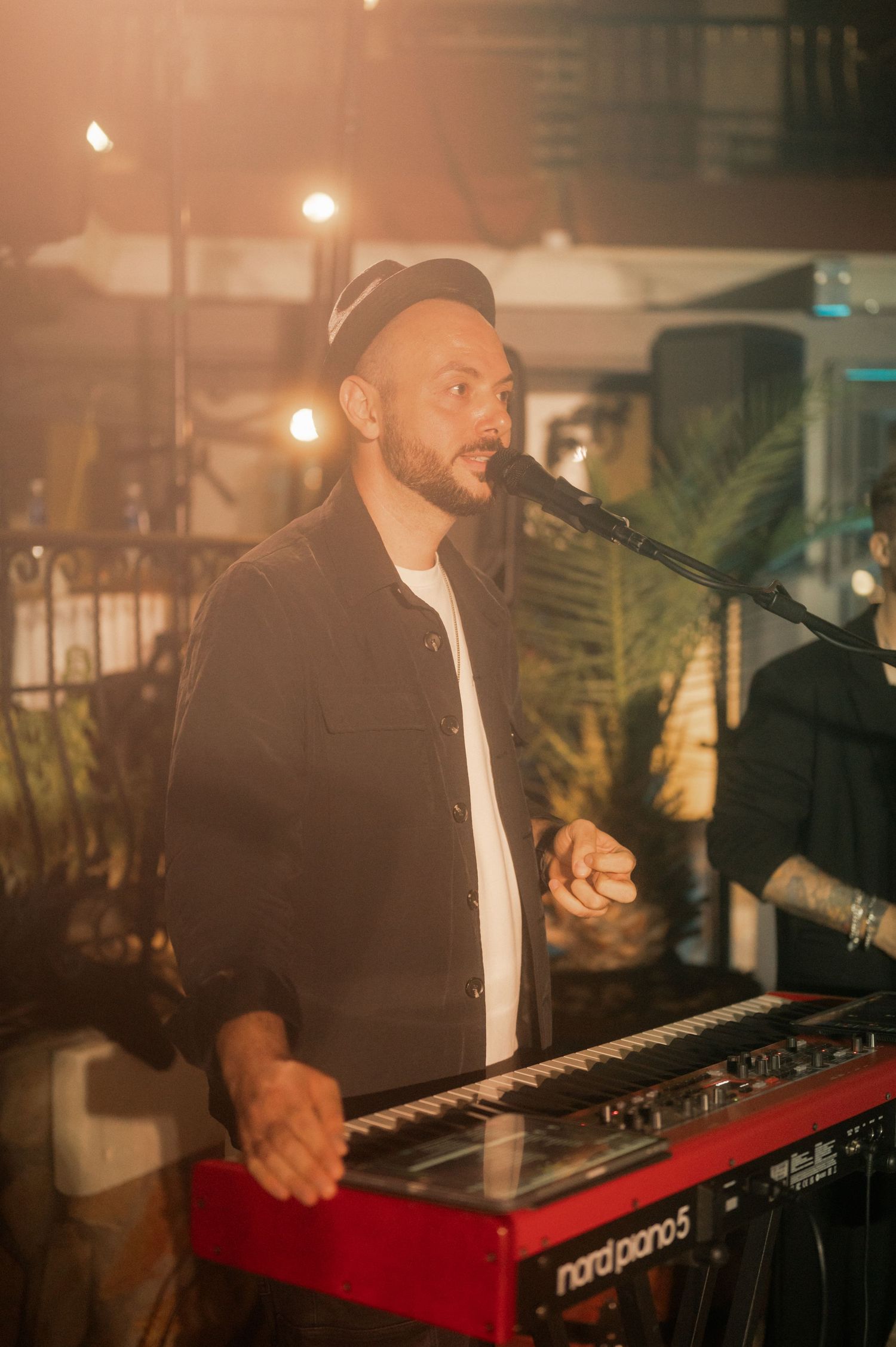 Musician performs on stage with a red Nord keyboard in warm ambient lighting with palm plants in the background.