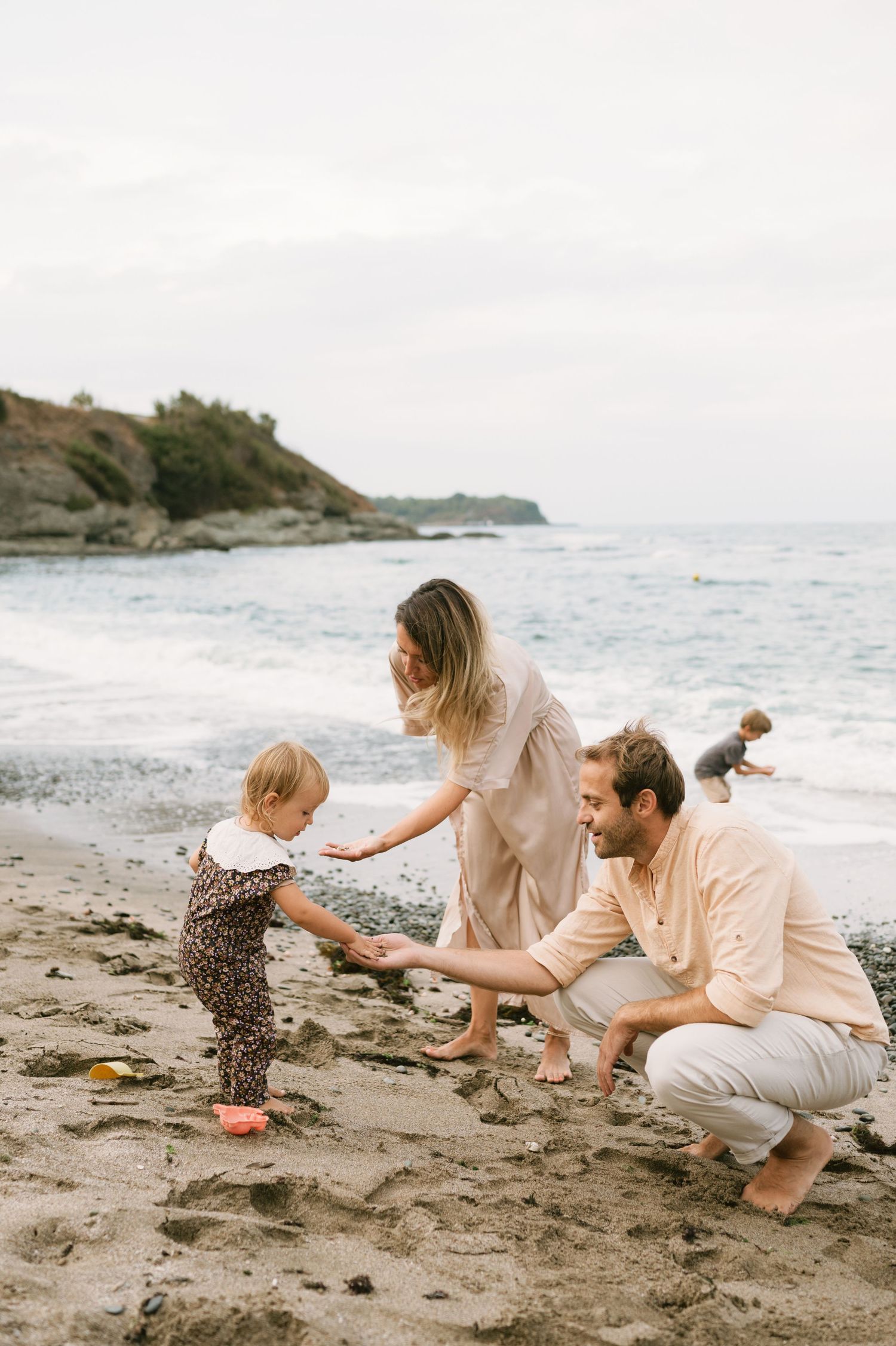 Family with toddler playing on a sandy beach near ocean waves, parents crouching down as child reaches out.