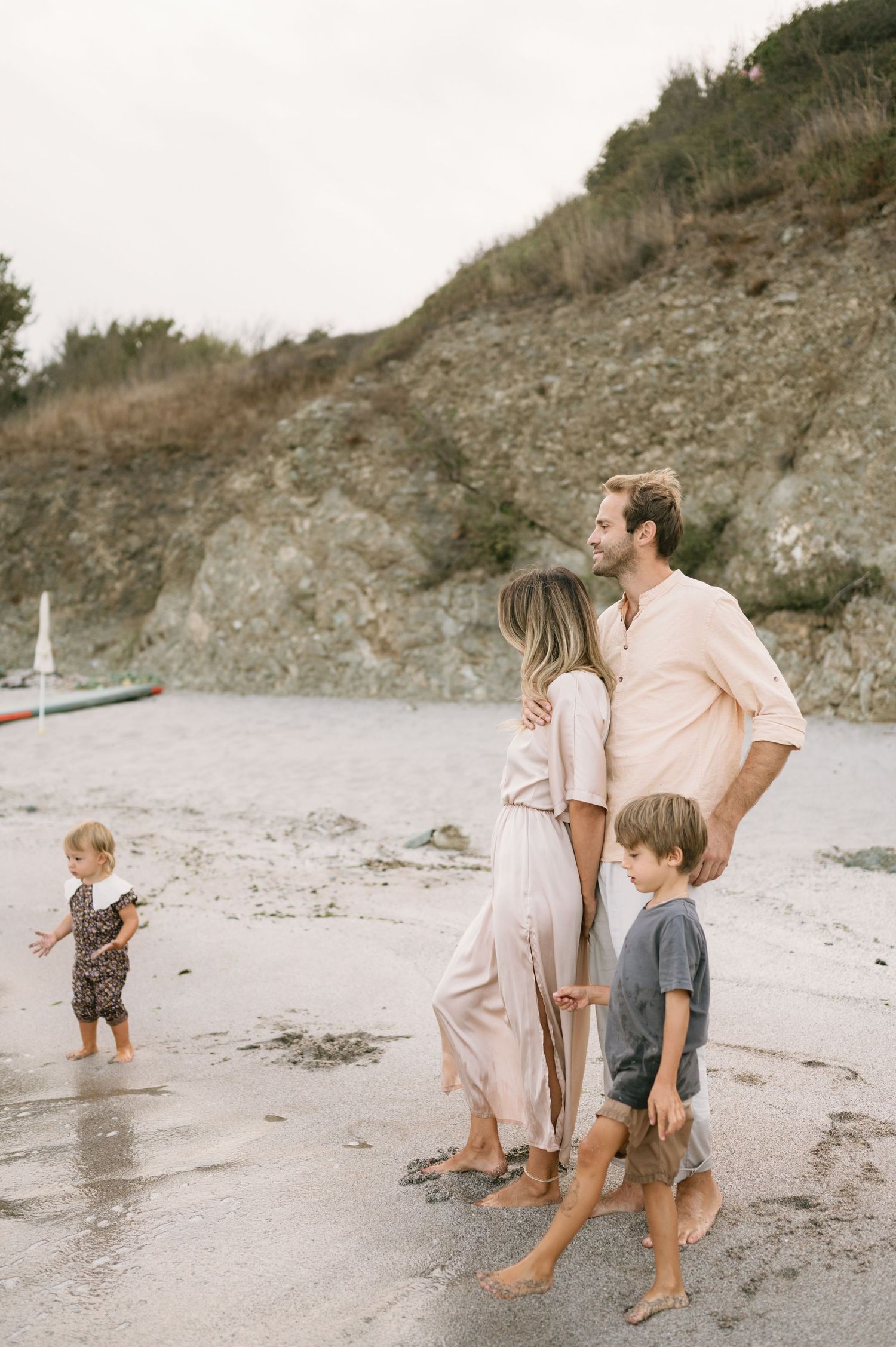 Family of four enjoying a peaceful beach day near rocky cliffs, parents embracing while children play in the sand.