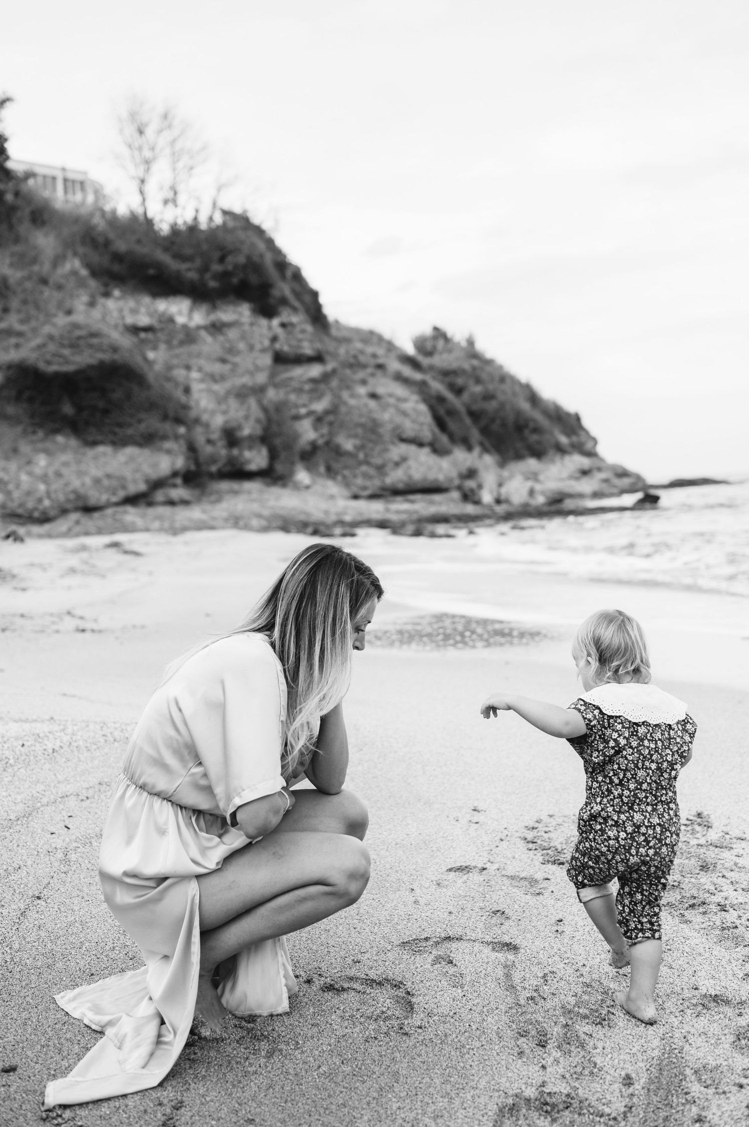 Black and white photo of a mother crouching on a sandy beach with her toddler pointing toward the ocean.