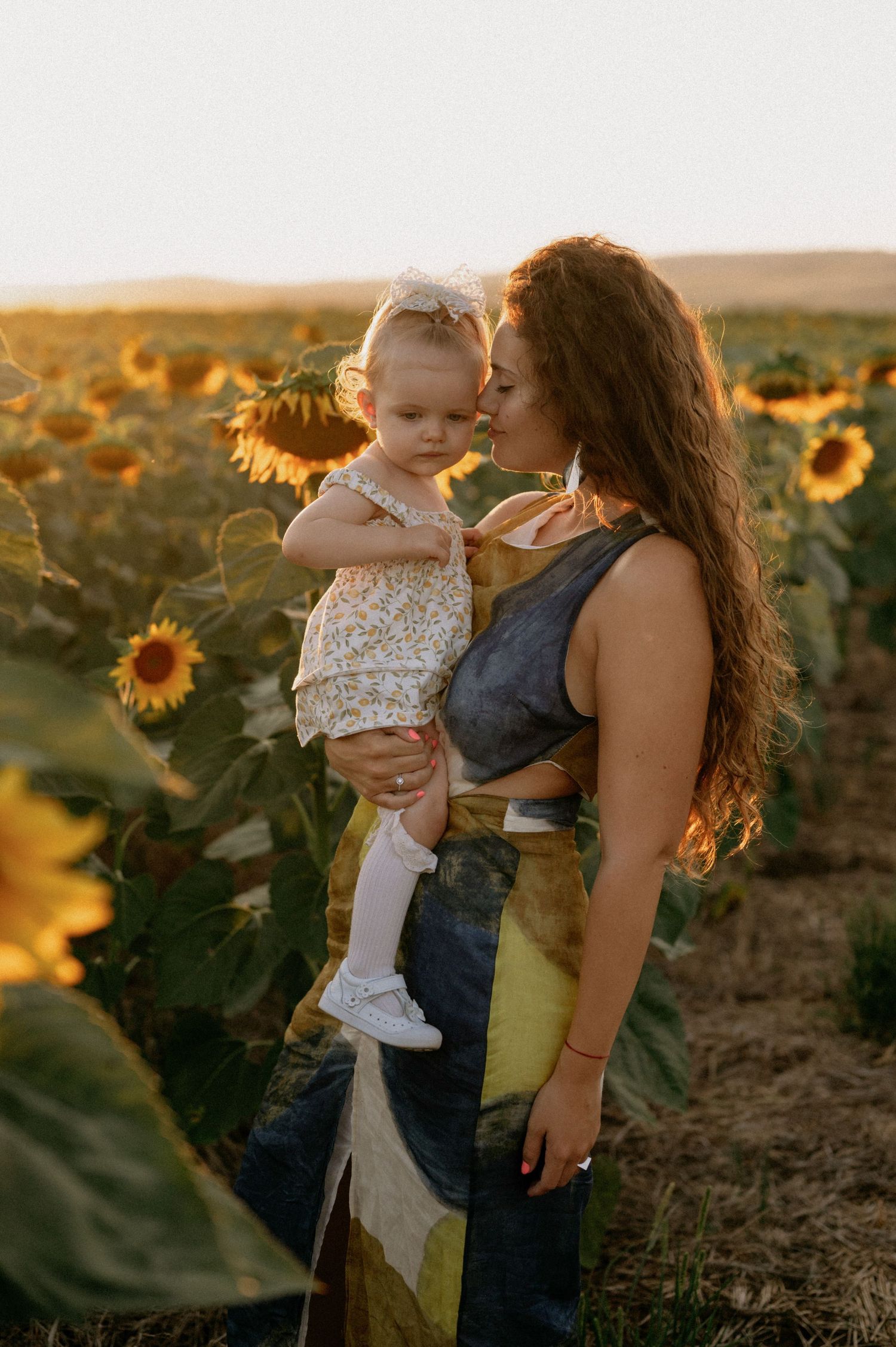 A mother kisses her toddler daughter in a golden sunflower field at sunset.