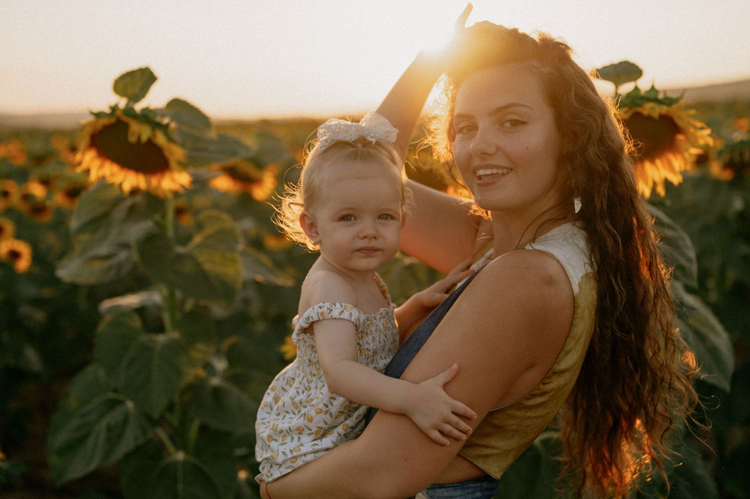 A young mother holds her baby girl in a sunflower field at golden hour sunset.