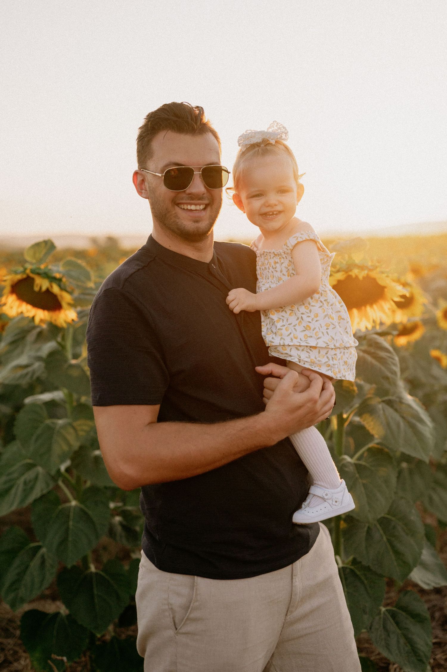 A smiling man in sunglasses holds a happy toddler girl in a floral dress in a sunflower field at golden hour.