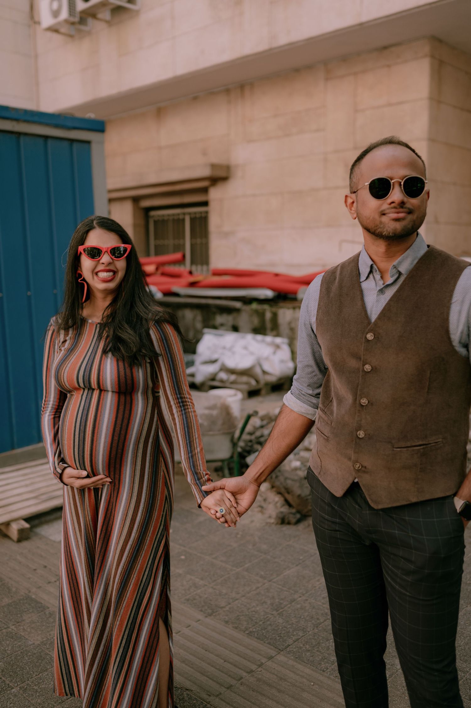Pregnant woman in striped dress holds hands with man in brown vest, both wearing sunglasses outdoors.