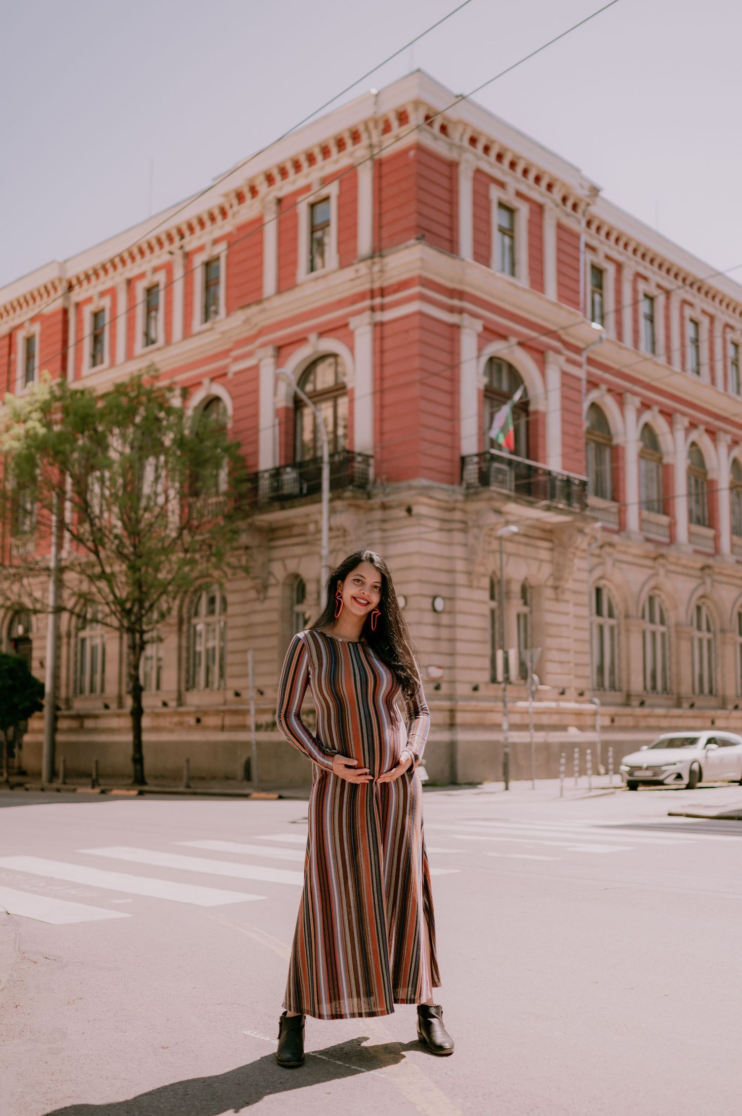 Woman in striped maxi dress smiling on sunny street corner near red historic building.
