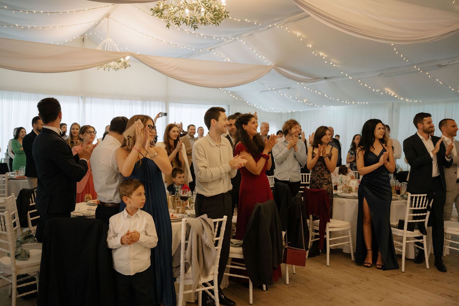 Guests applauding and celebrating at an elegant wedding reception in a white draped marquee tent.