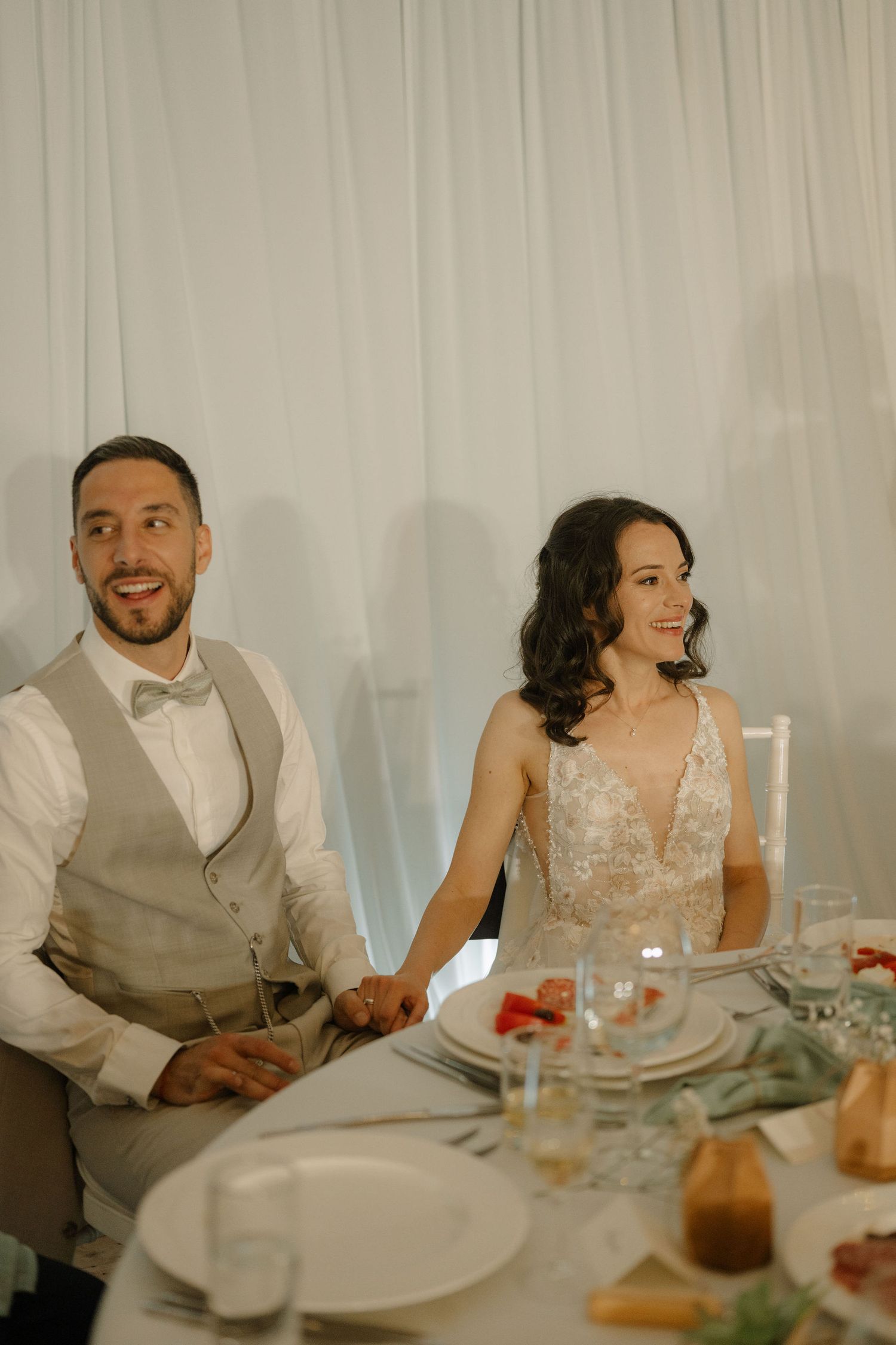 Bride and groom smiling at wedding reception table, groom in beige vest and bride in lace gown holding hands.