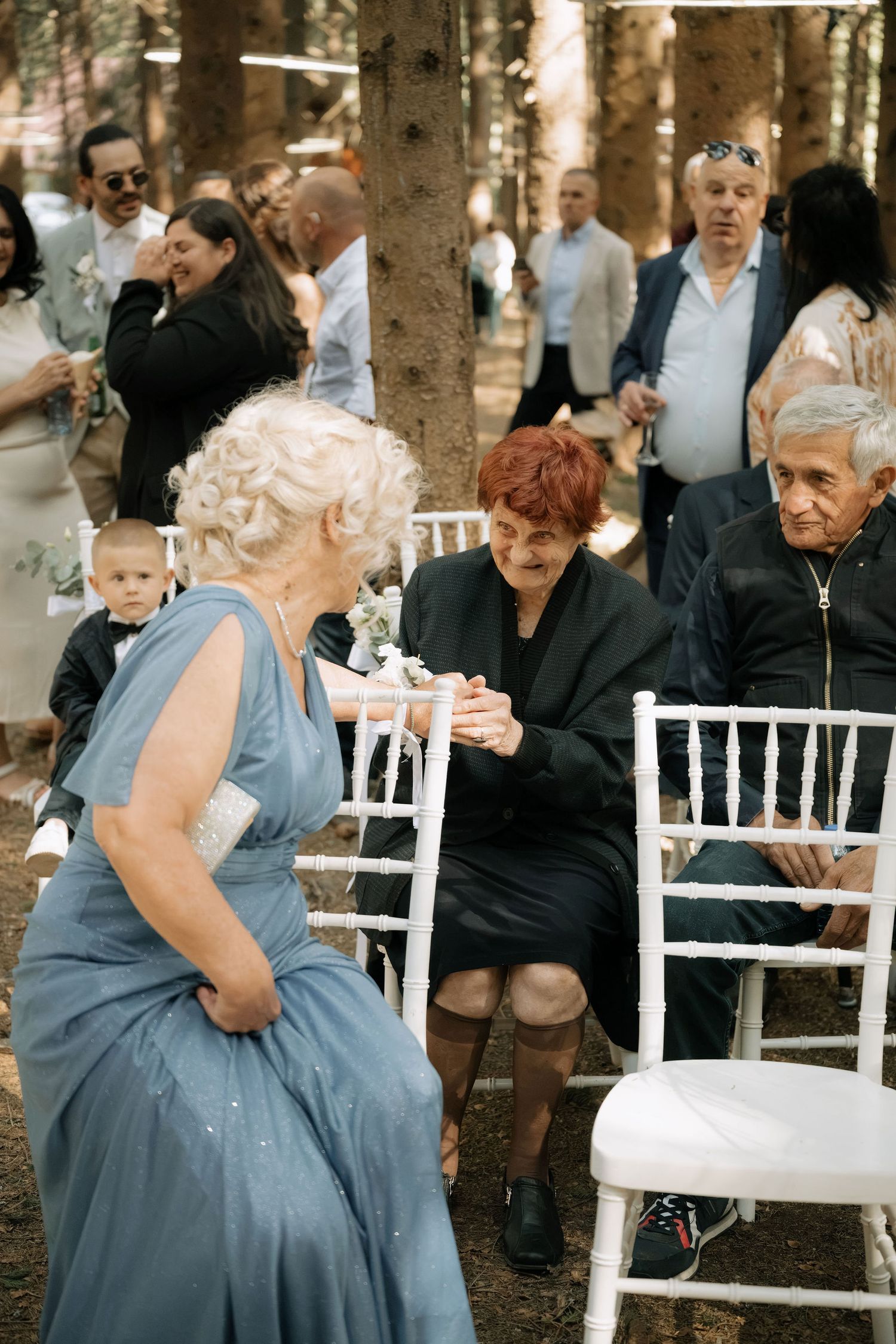 Elderly woman in blue gown interacts with seated guests at an outdoor woodland wedding ceremony.