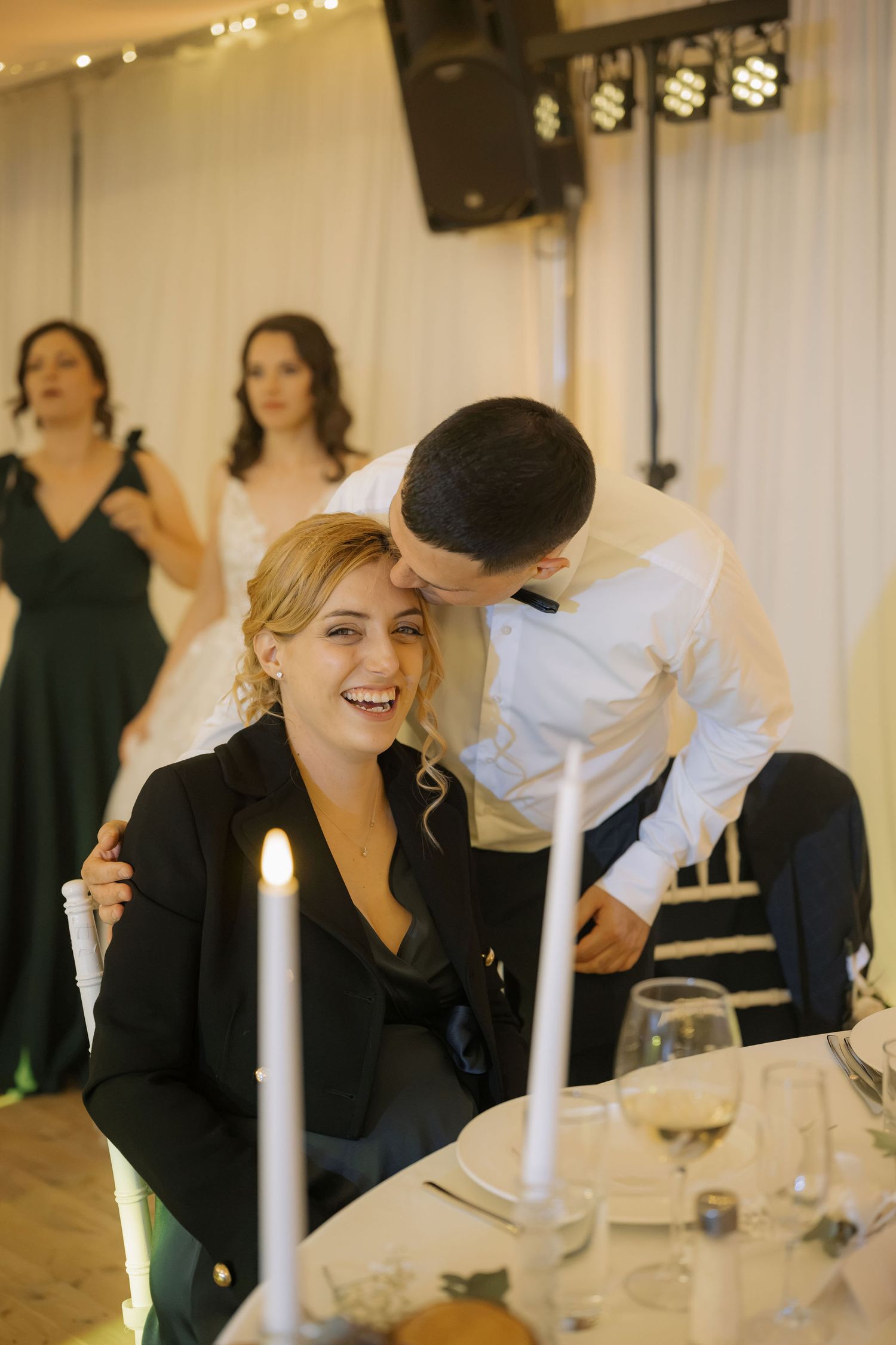 A laughing blonde woman in a black blazer sits at a candlelit wedding reception table as a man leans in to whisper.