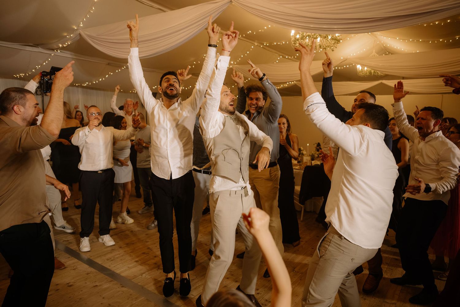 Joyful wedding reception dance floor with guests celebrating, arms raised, string lights overhead.