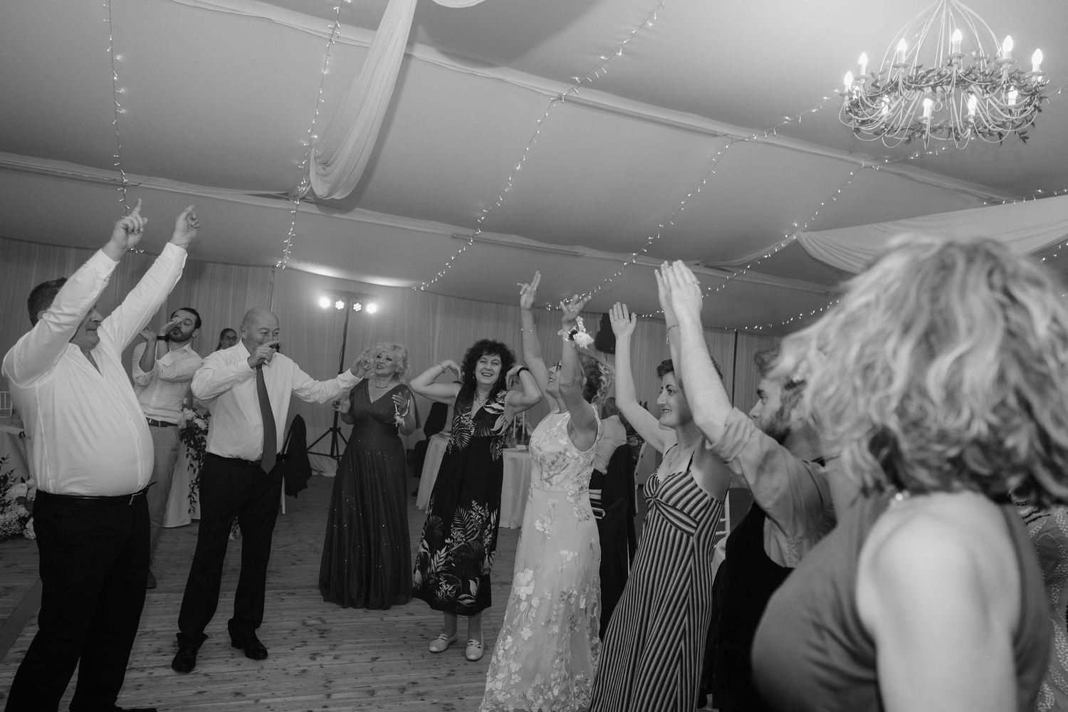 Guests dancing joyfully at a wedding reception in a decorated ballroom, black and white photo.