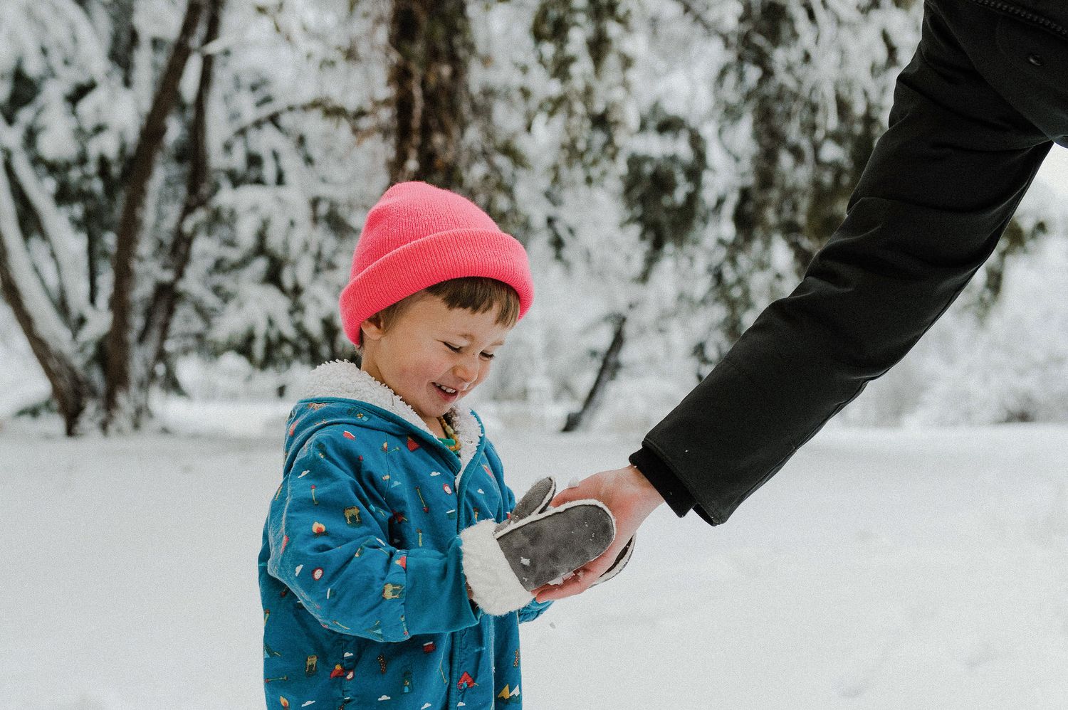 Child in blue winter coat plays in snowy forest while adult helps from behind.