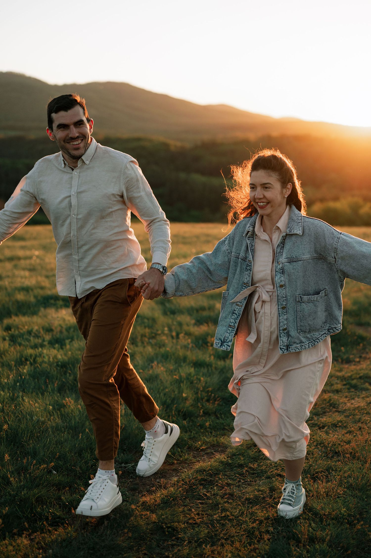 A couple runs playfully through a grassy field at sunset, wearing casual outfits and white sneakers.