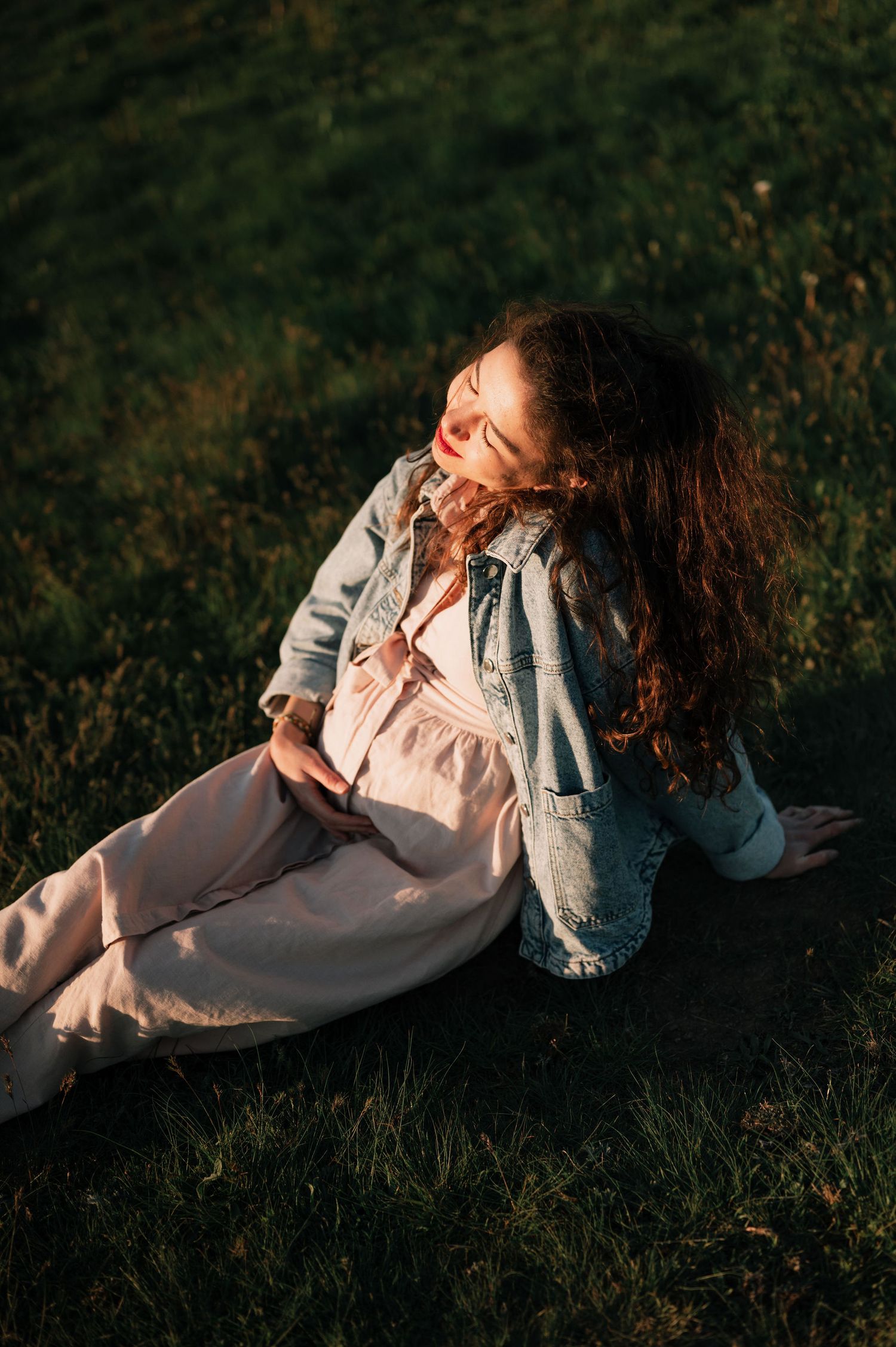 A person in casual attire sits on grass during golden hour, with curly hair illuminated by warm sunlight.