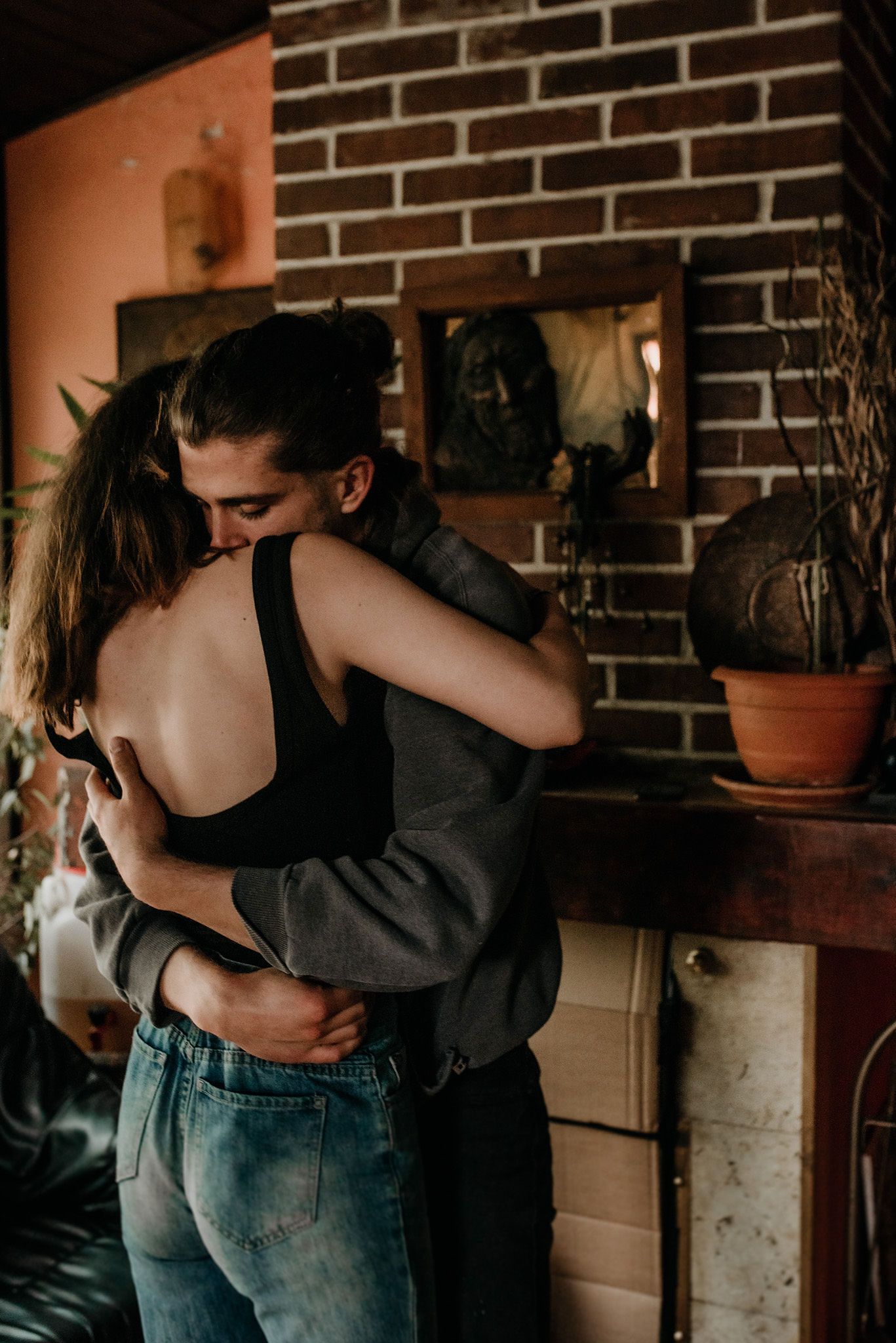 A couple embraces intimately in a rustic kitchen setting with brick walls and warm lighting.