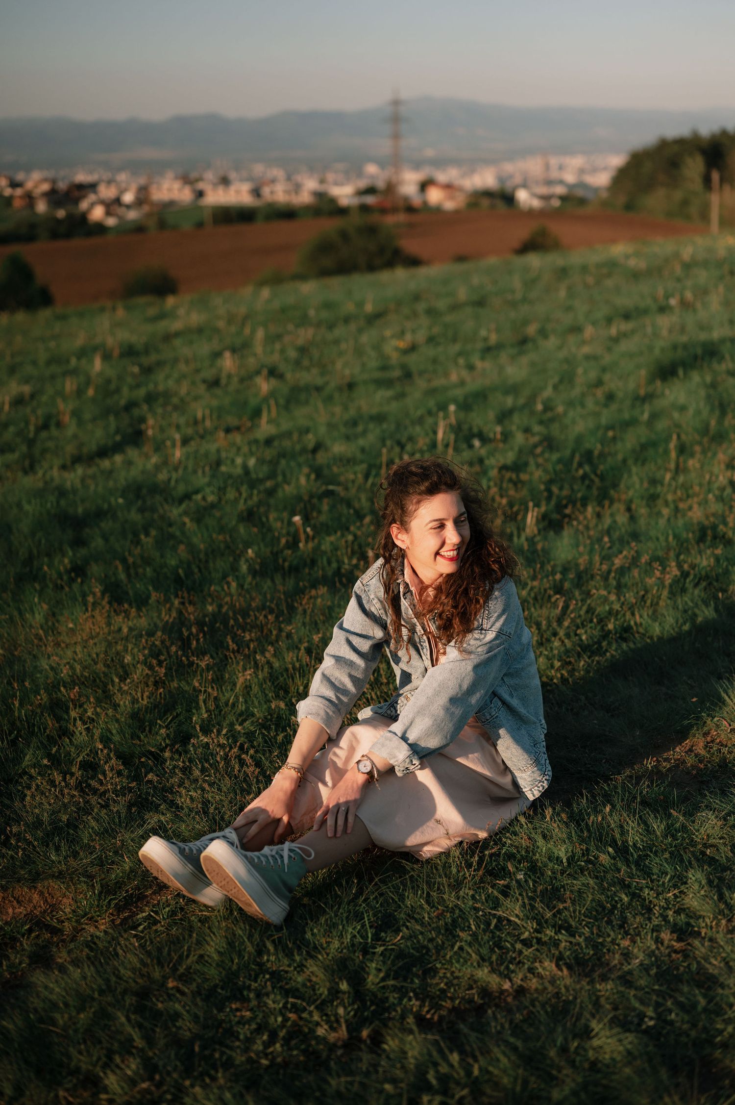 Person relaxing on grass with cityscape and mountains visible in background at sunset.