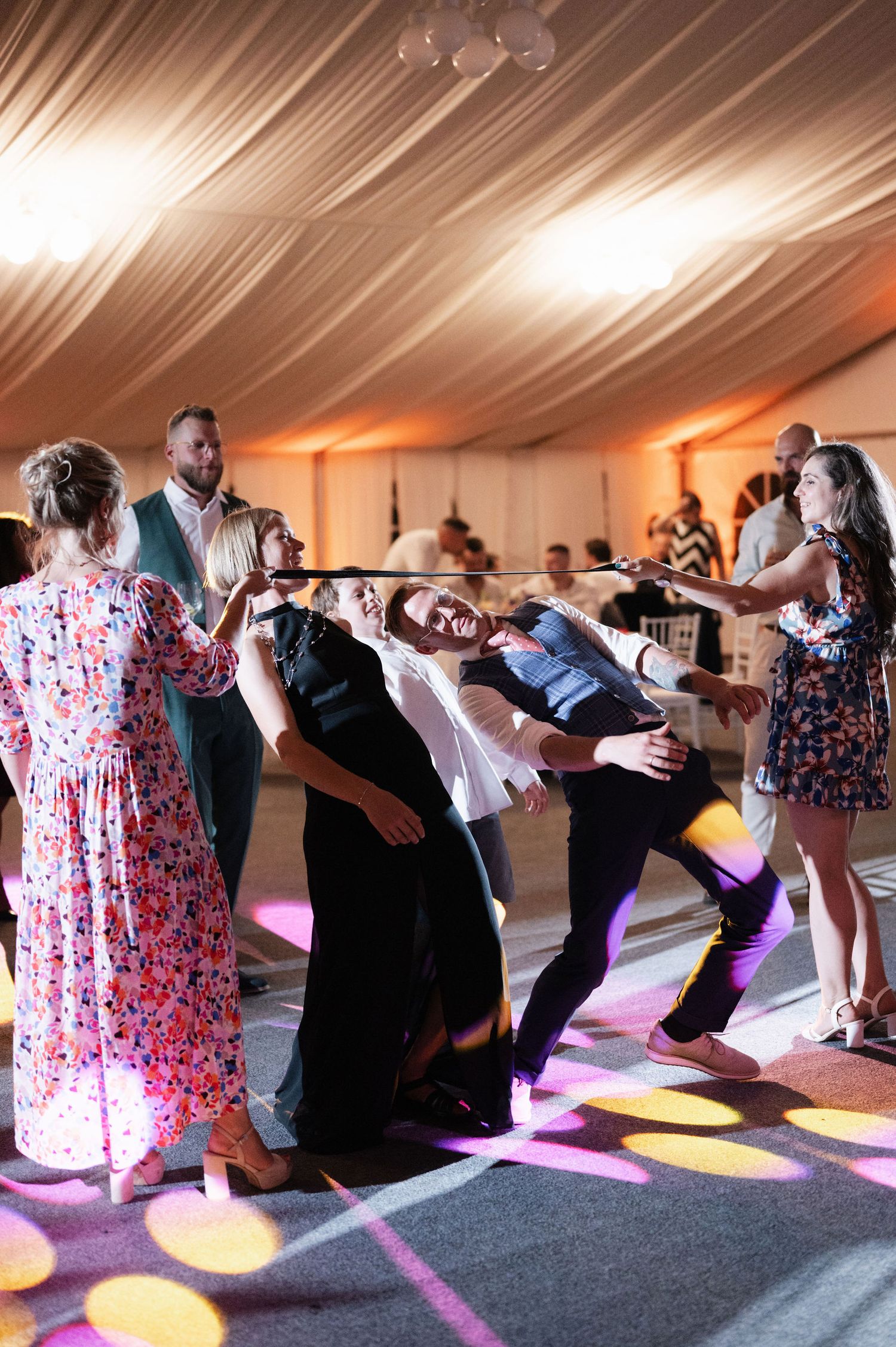 Wedding guests dance and lean back in a synchronized line dance formation under a white tent with colorful lighting.