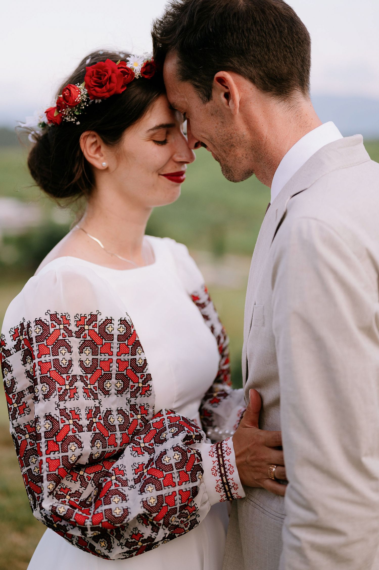 Wedding couple embraces on grassy hill wearing white dress with red embroidery and beige suit.