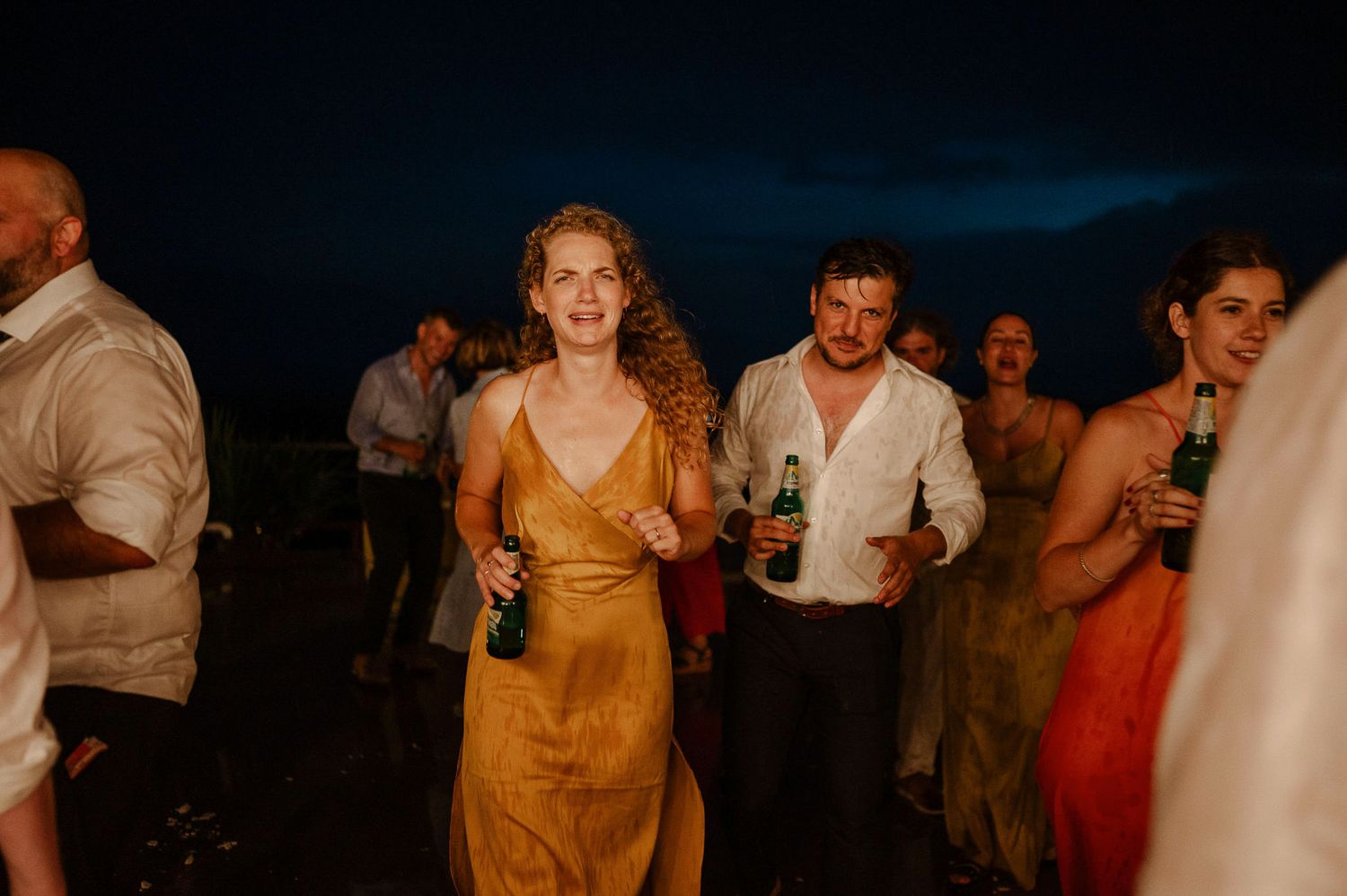 Wedding celebration at night with guests mingling and dancing under outdoor lighting.