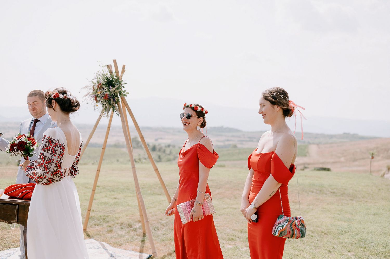 Wedding party in red dresses and white attire gather on hillside during outdoor ceremony.