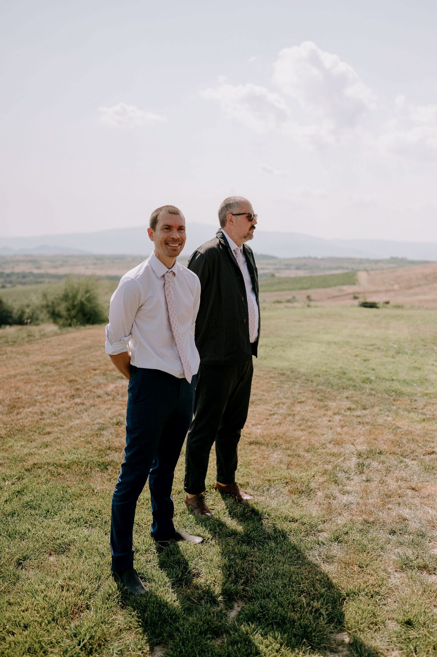 Two people standing together on a grassy hillside overlooking a scenic landscape.