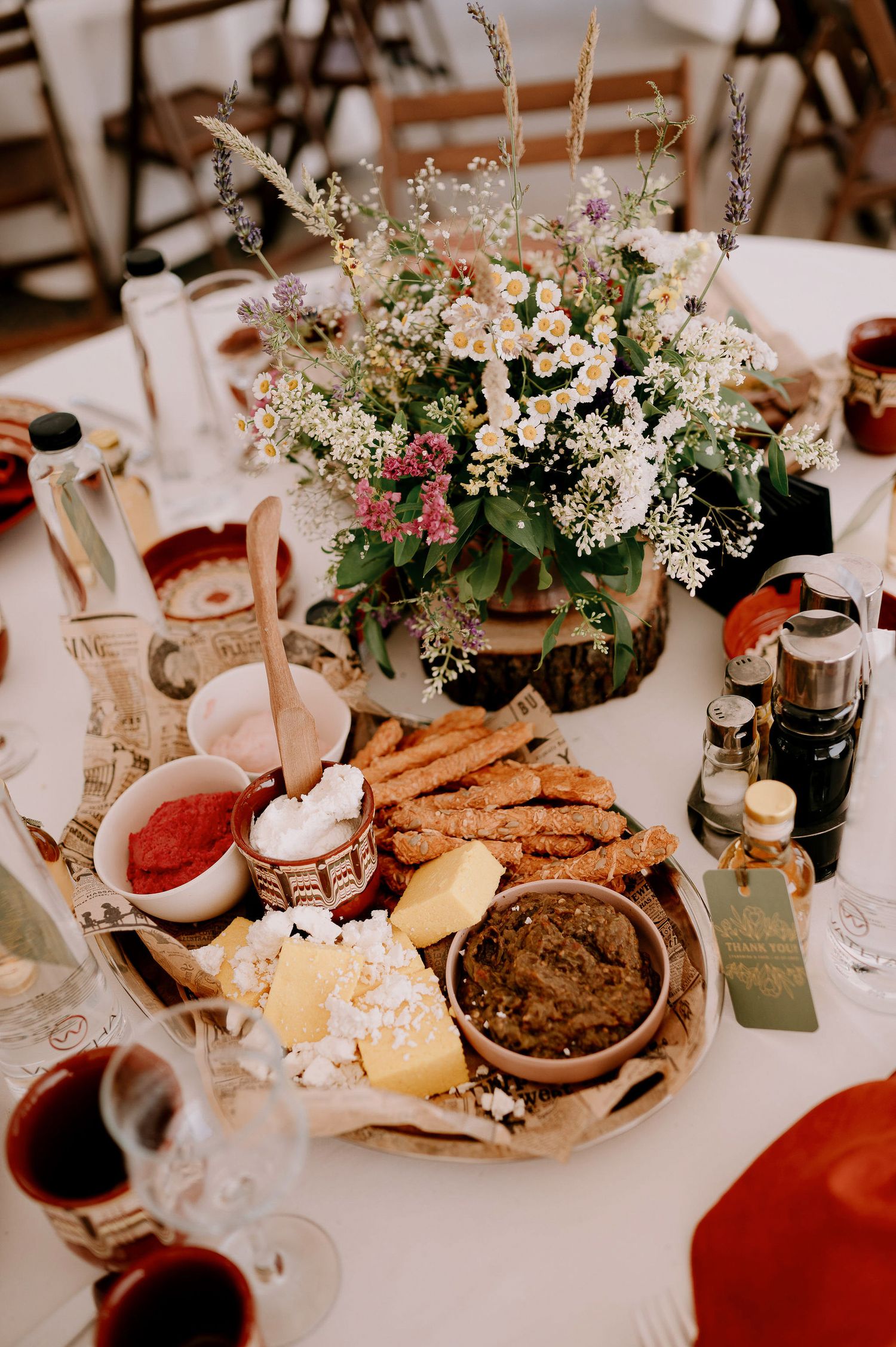 Rustic wedding table setting with floral centerpiece, appetizer platter, and wine glasses on white tablecloth.