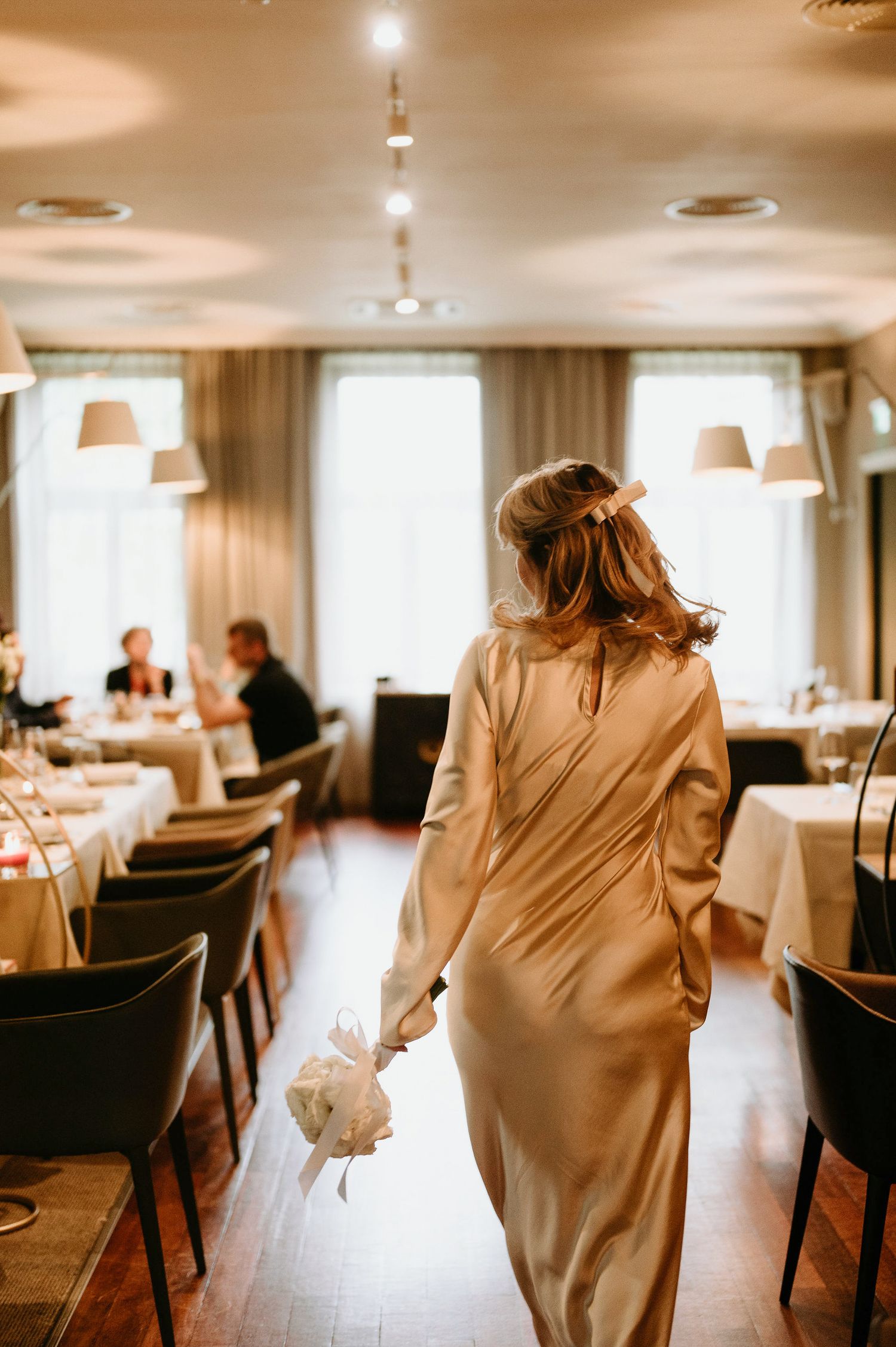 A person in a cream-colored dress walks through an elegant dining room with natural lighting.