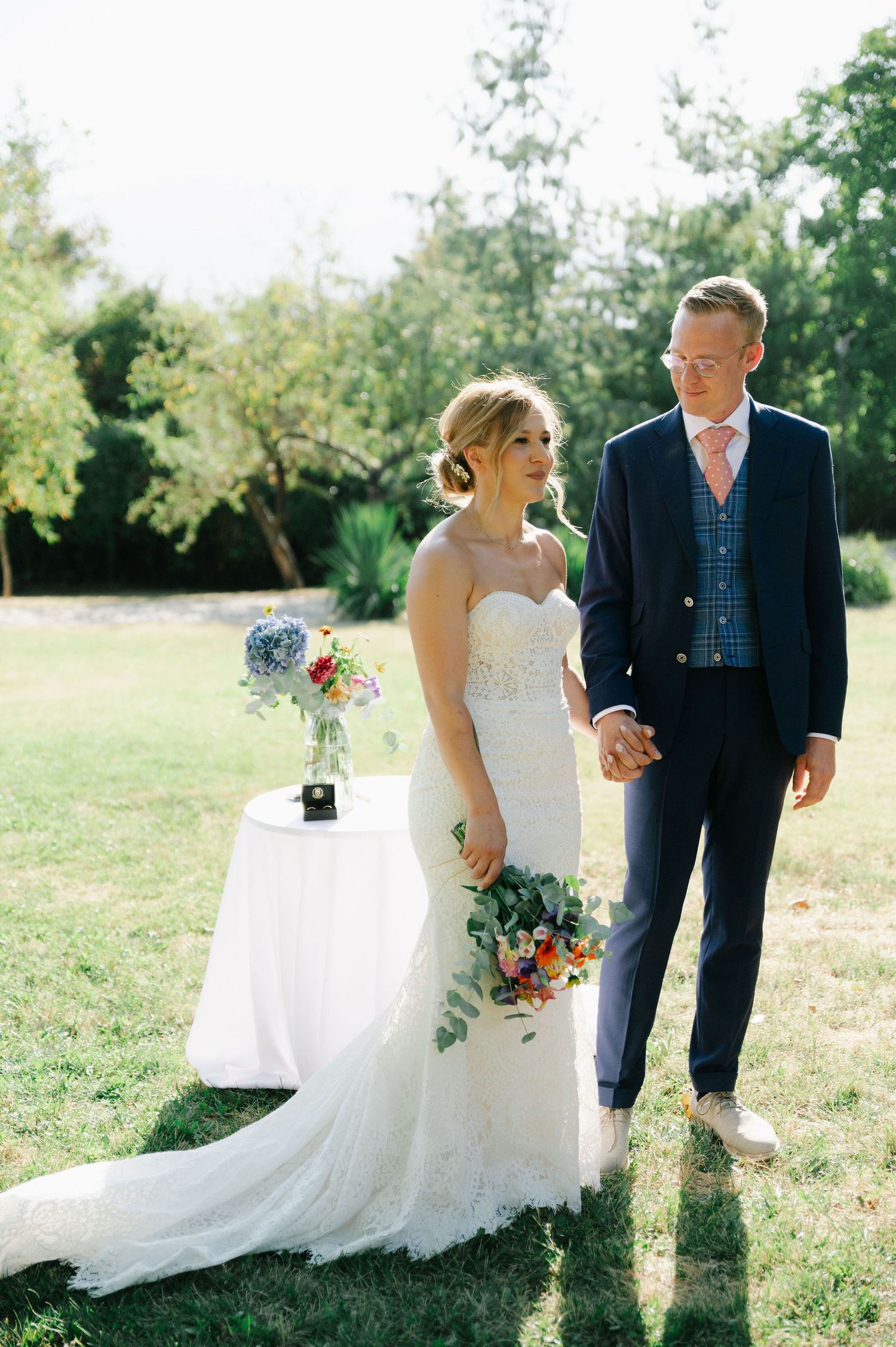 Bride in white gown and groom in navy suit pose together in a sunlit field.
