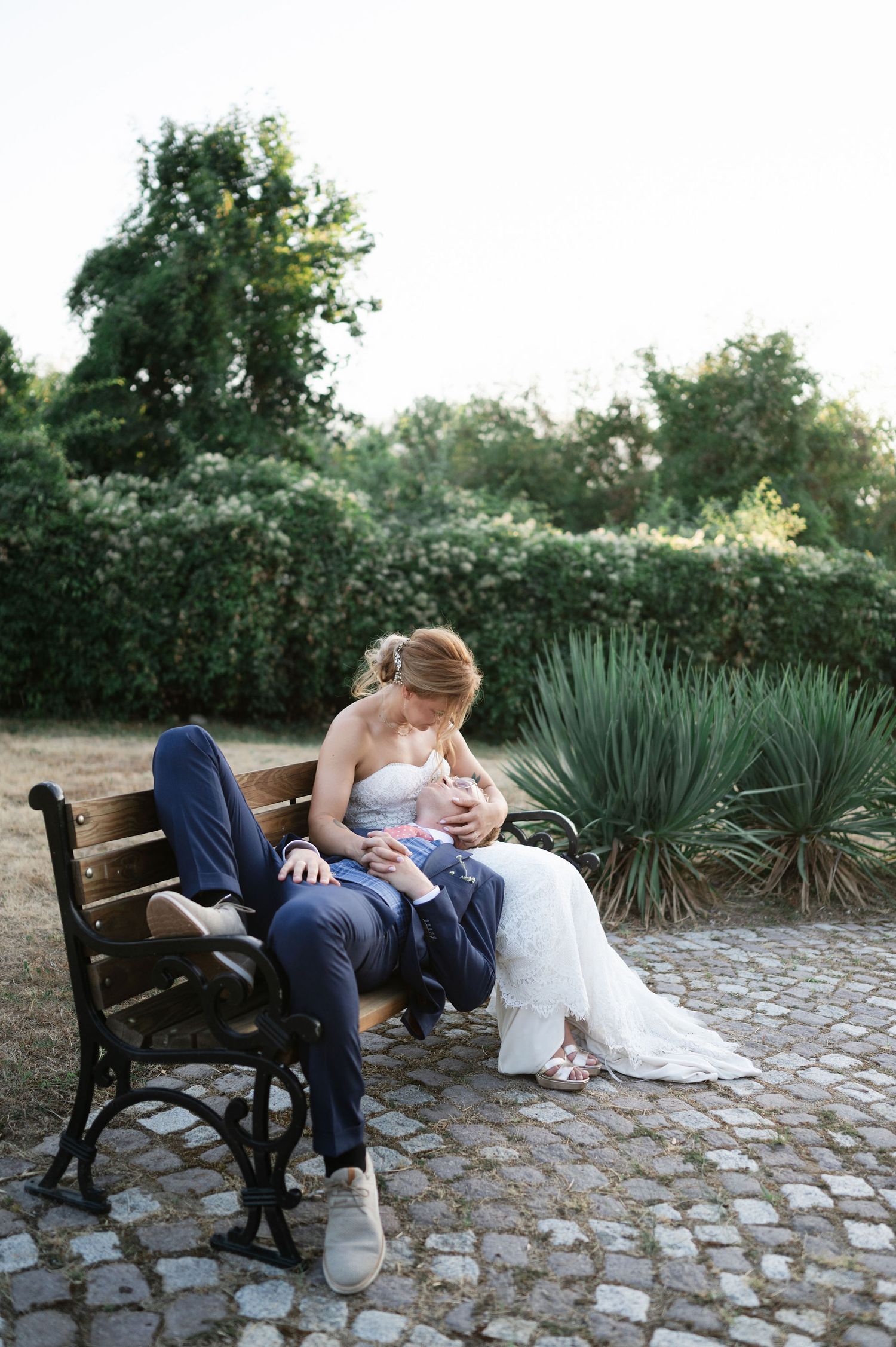 Romantic wedding photo series on a rustic bench with cobblestone ground and garden backdrop.
