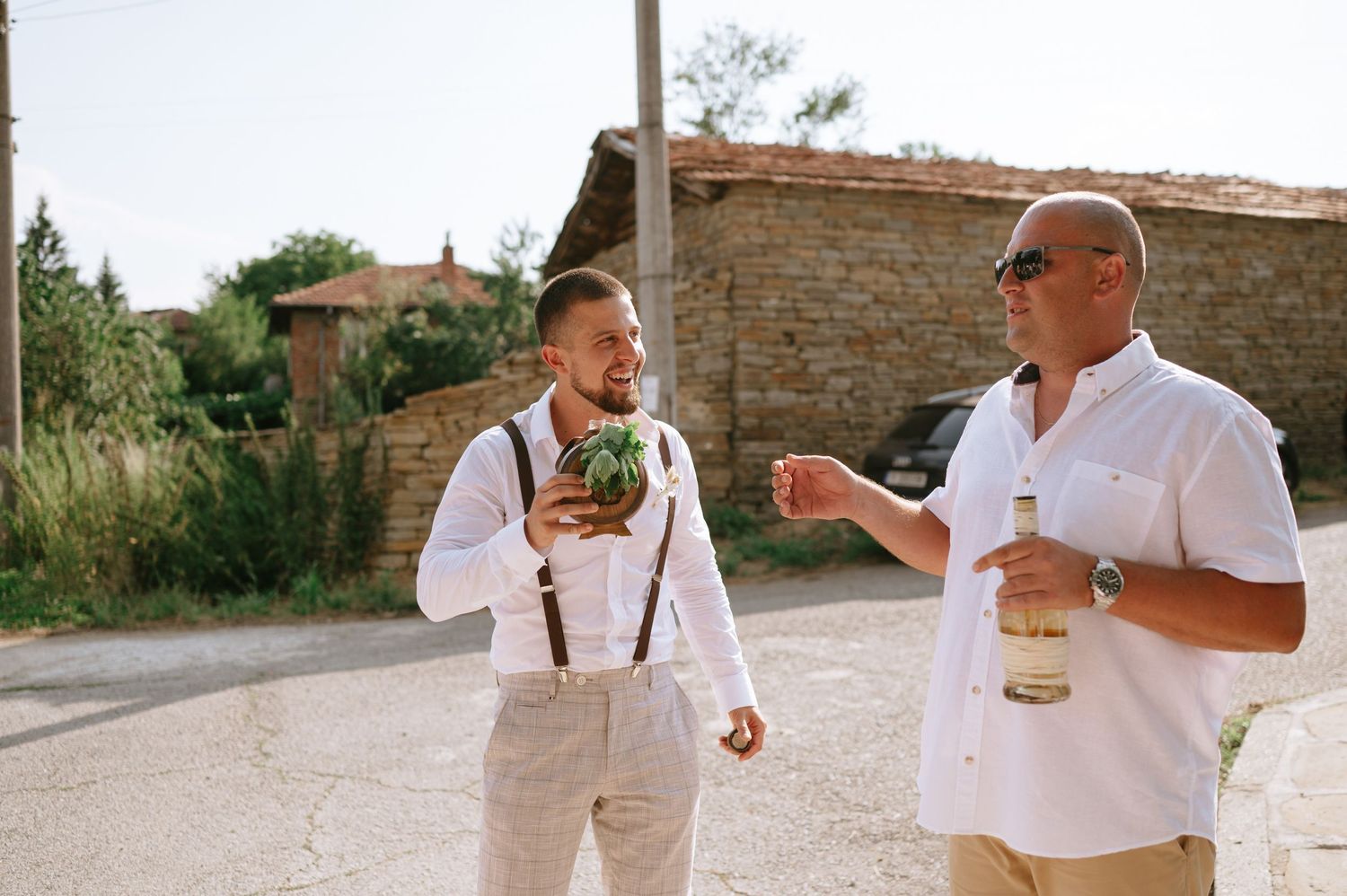 Groomsmen in light colored suspenders and white shirts sharing drinks outdoors.