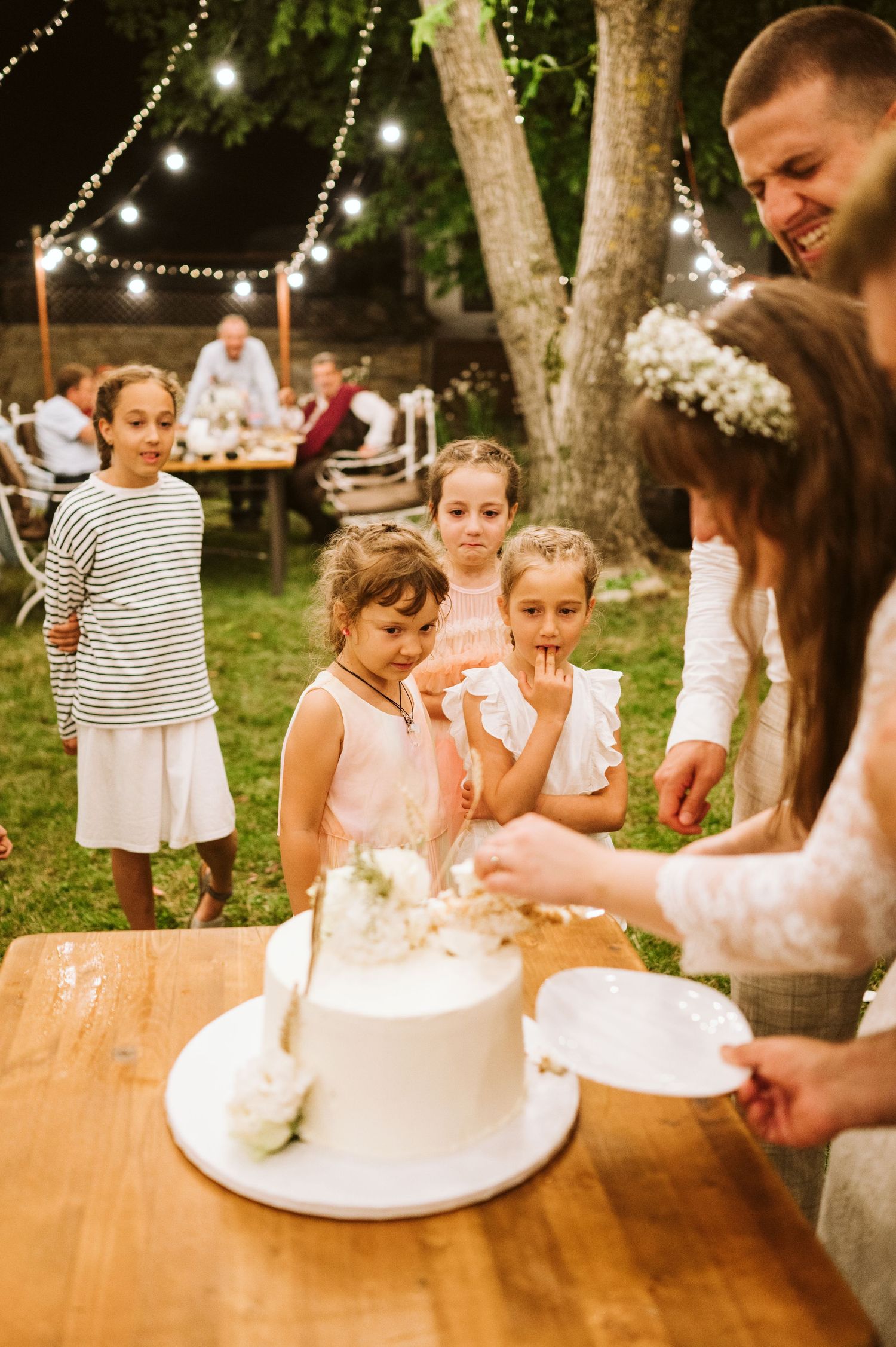 Children eagerly watch cake cutting at outdoor wedding reception with string lights.