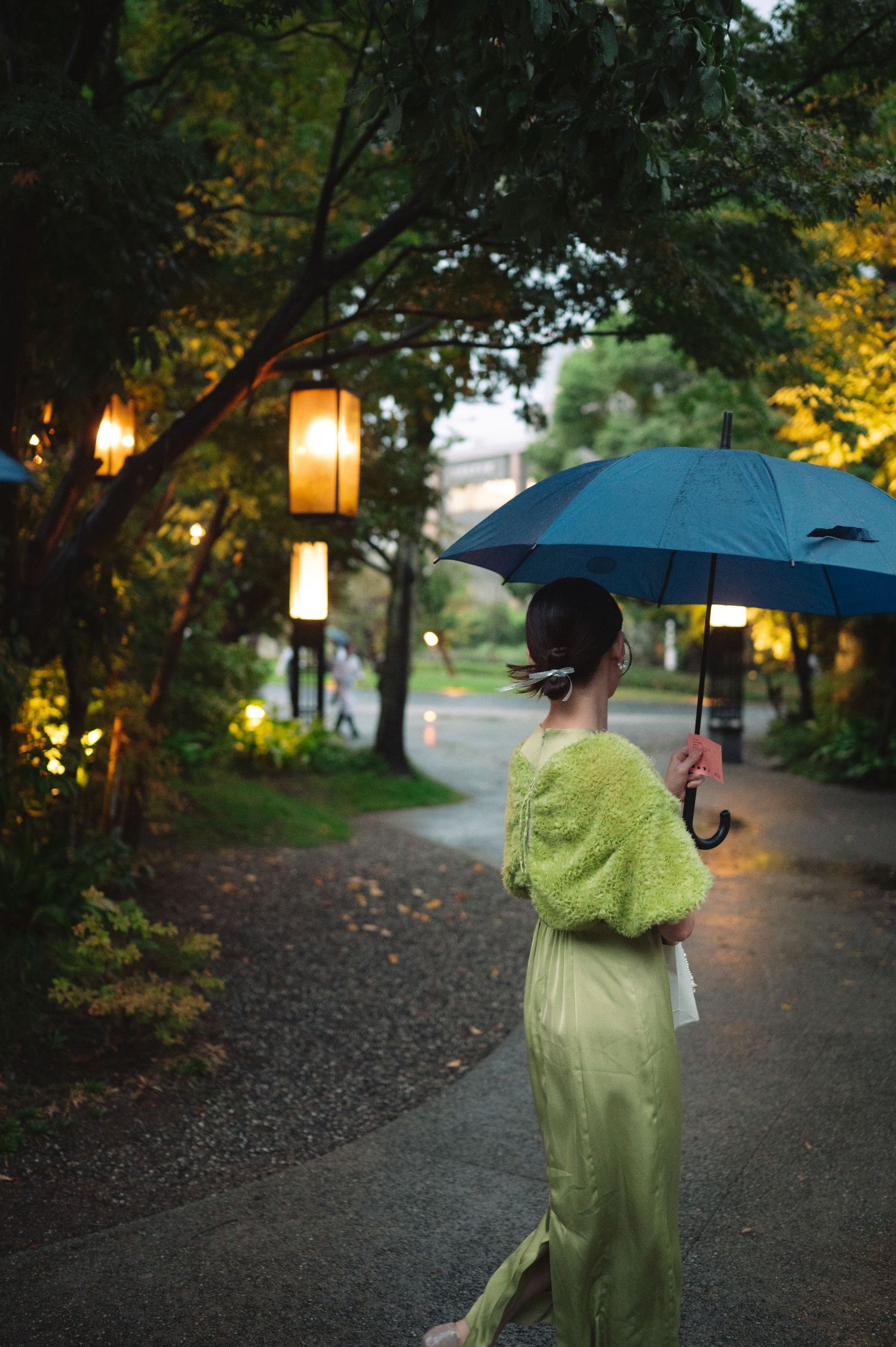 Person in rain gear walking with umbrella on illuminated garden path at twilight.