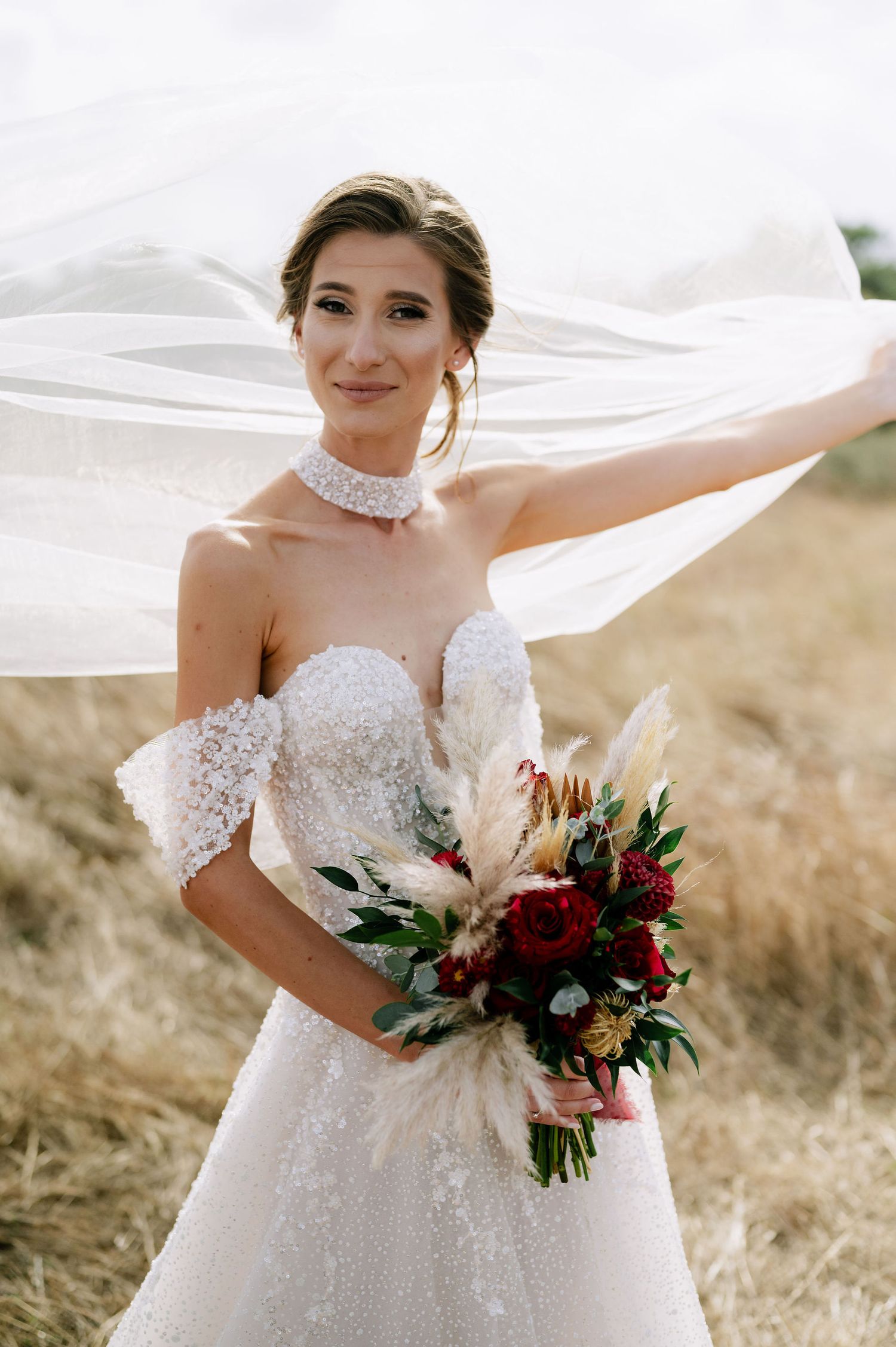A bride in a strapless beaded gown holds a red and white bouquet while her veil flows in the breeze.