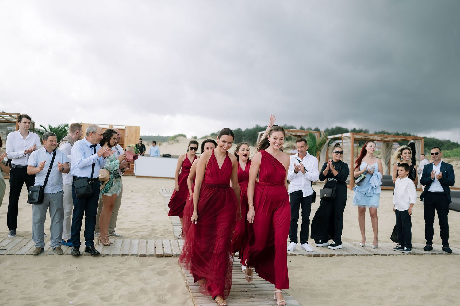 Wedding party in burgundy dresses poses on beach with cloudy sky backdrop.