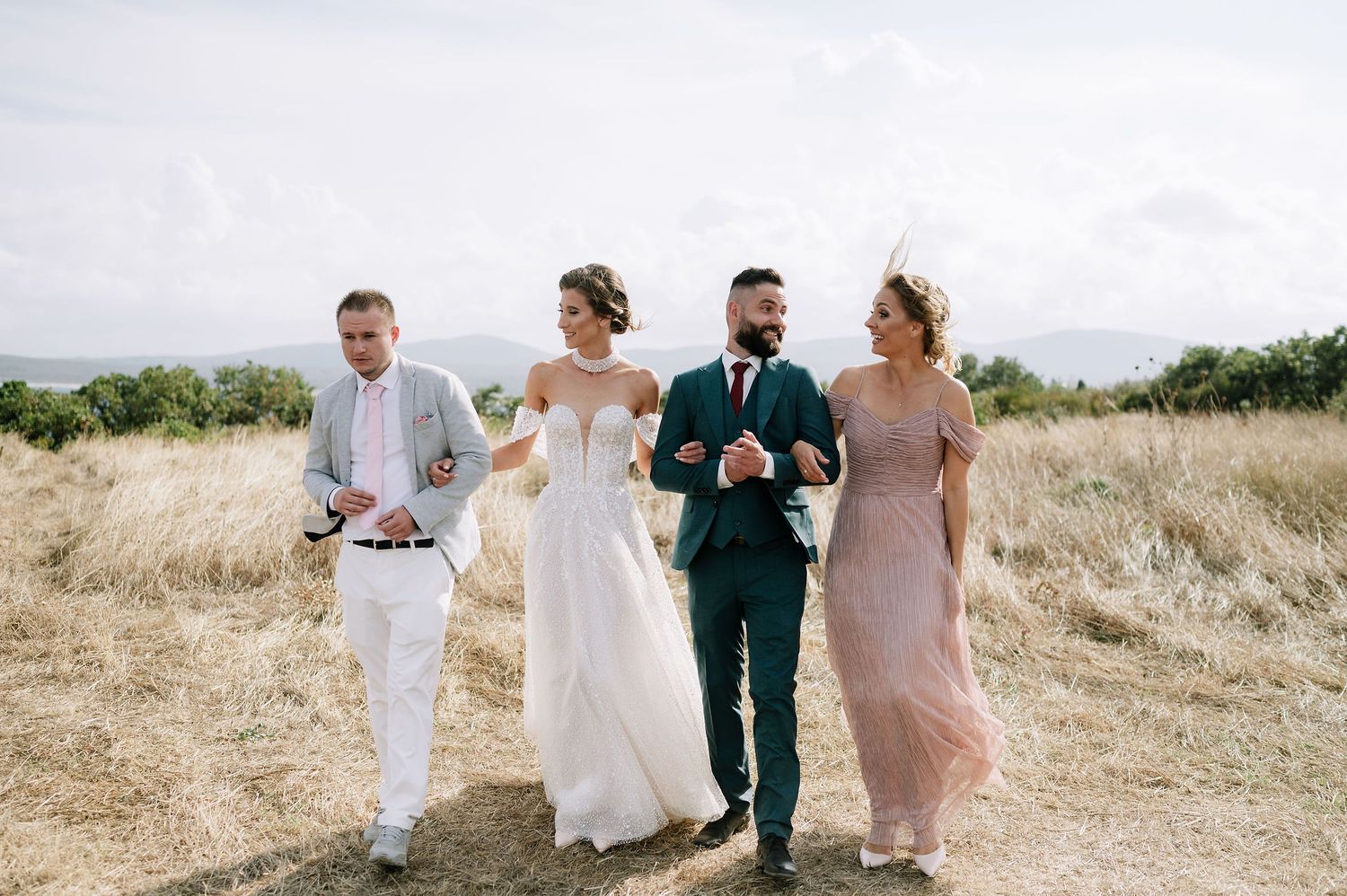 Wedding party walking together through a sun-drenched field in formal attire.