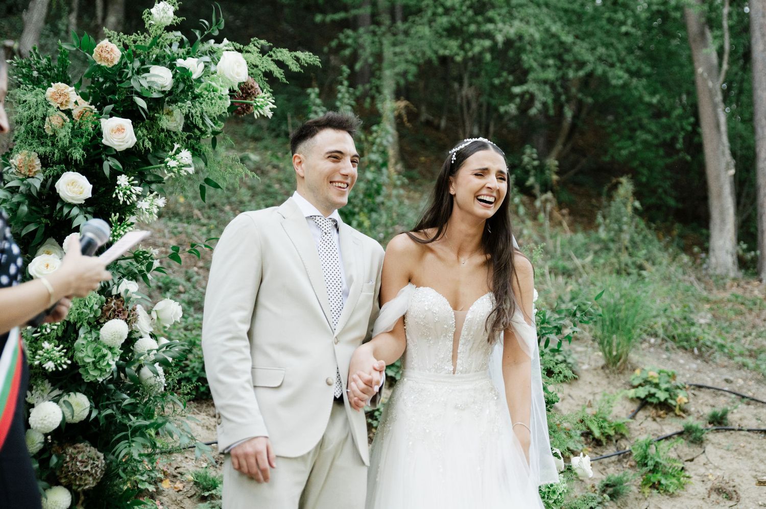 Joyful wedding moment captured under an arch of cream and blush roses in woodland setting.