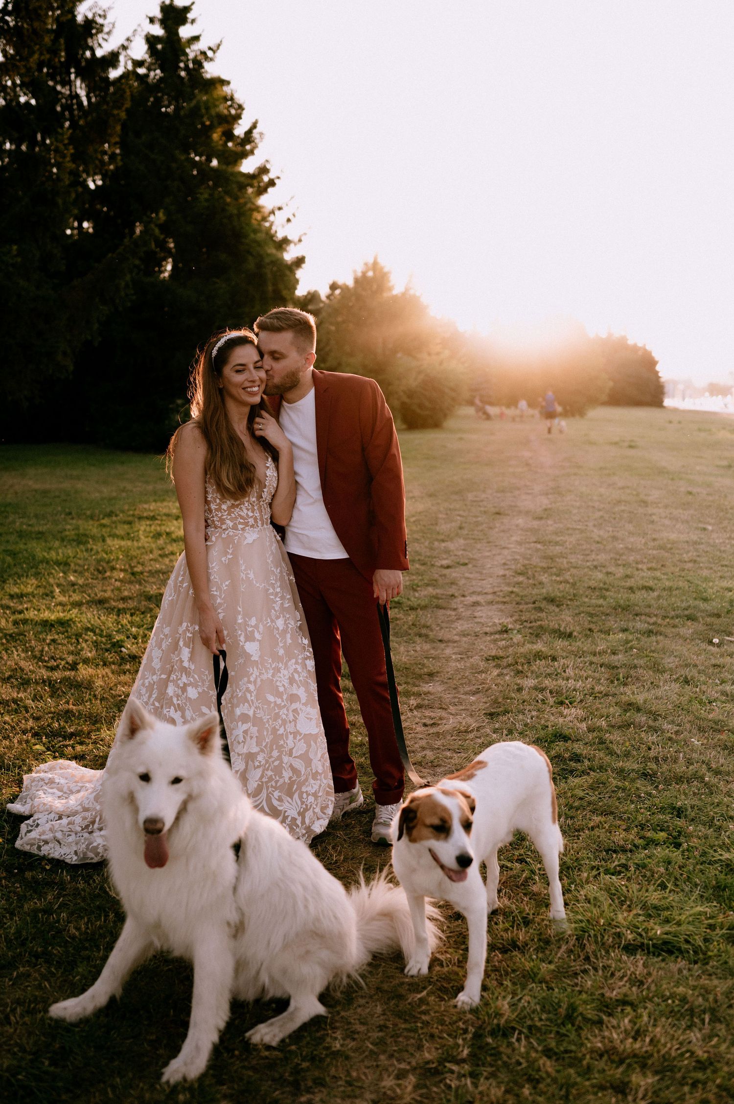 Newlyweds in burgundy and white pose with their white dogs during golden hour.