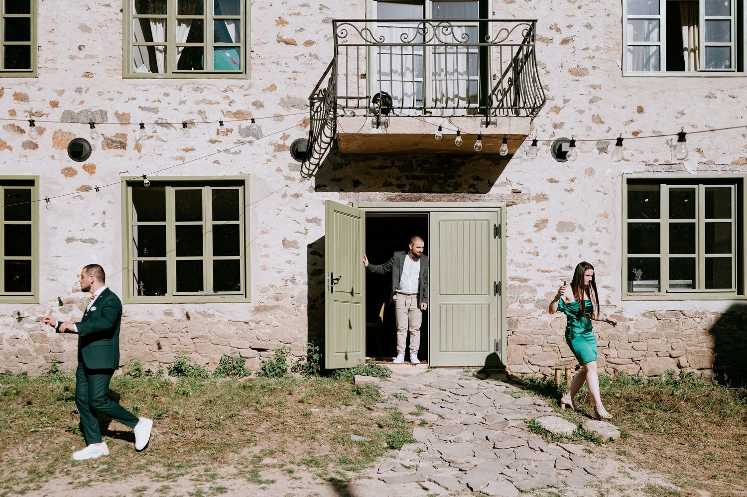 Wedding guests gather outside a rustic stone building with white trim windows during an outdoor celebration.